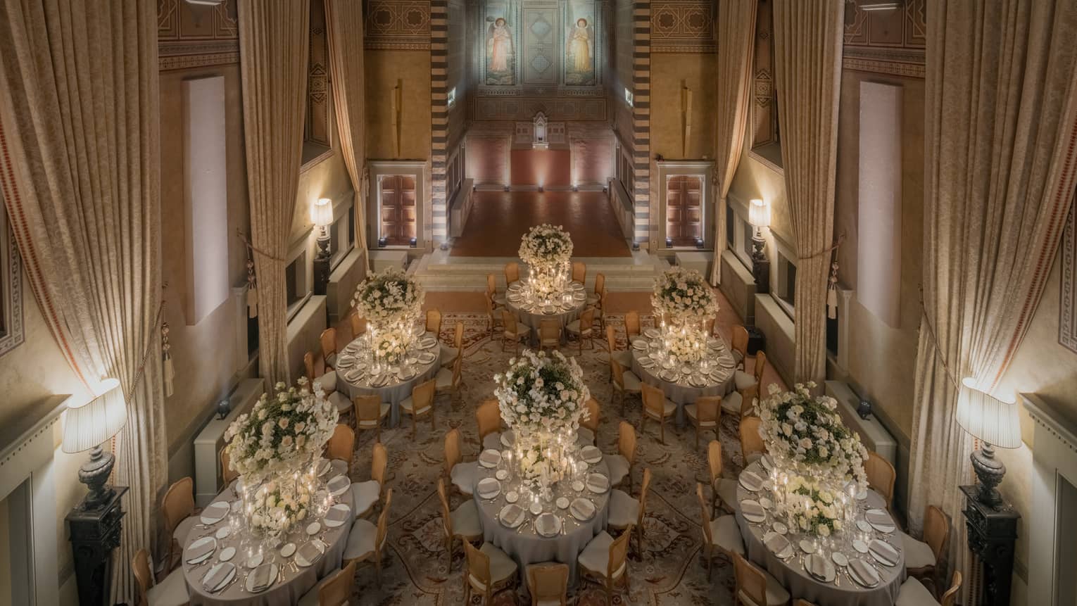 A large softly lit event space with high ceilings, drapes, recessed and table lamp lighting, and round tables with chairs, place settings and floral centrepieces