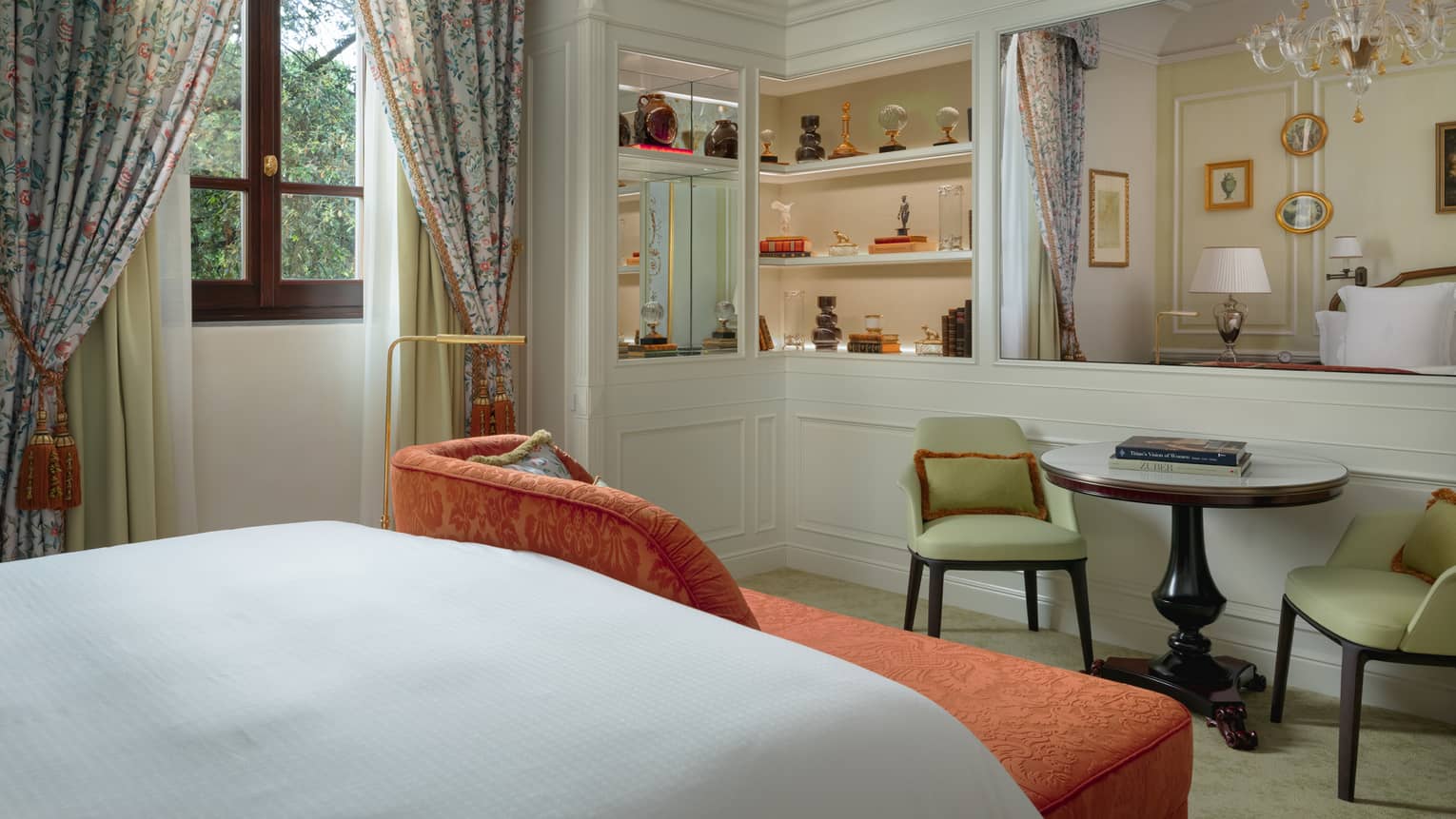 Elegant hotel room featuring a king bed, orange chaise at the foot of the bed, a table and two light green chairs, and a window with patterned curtains