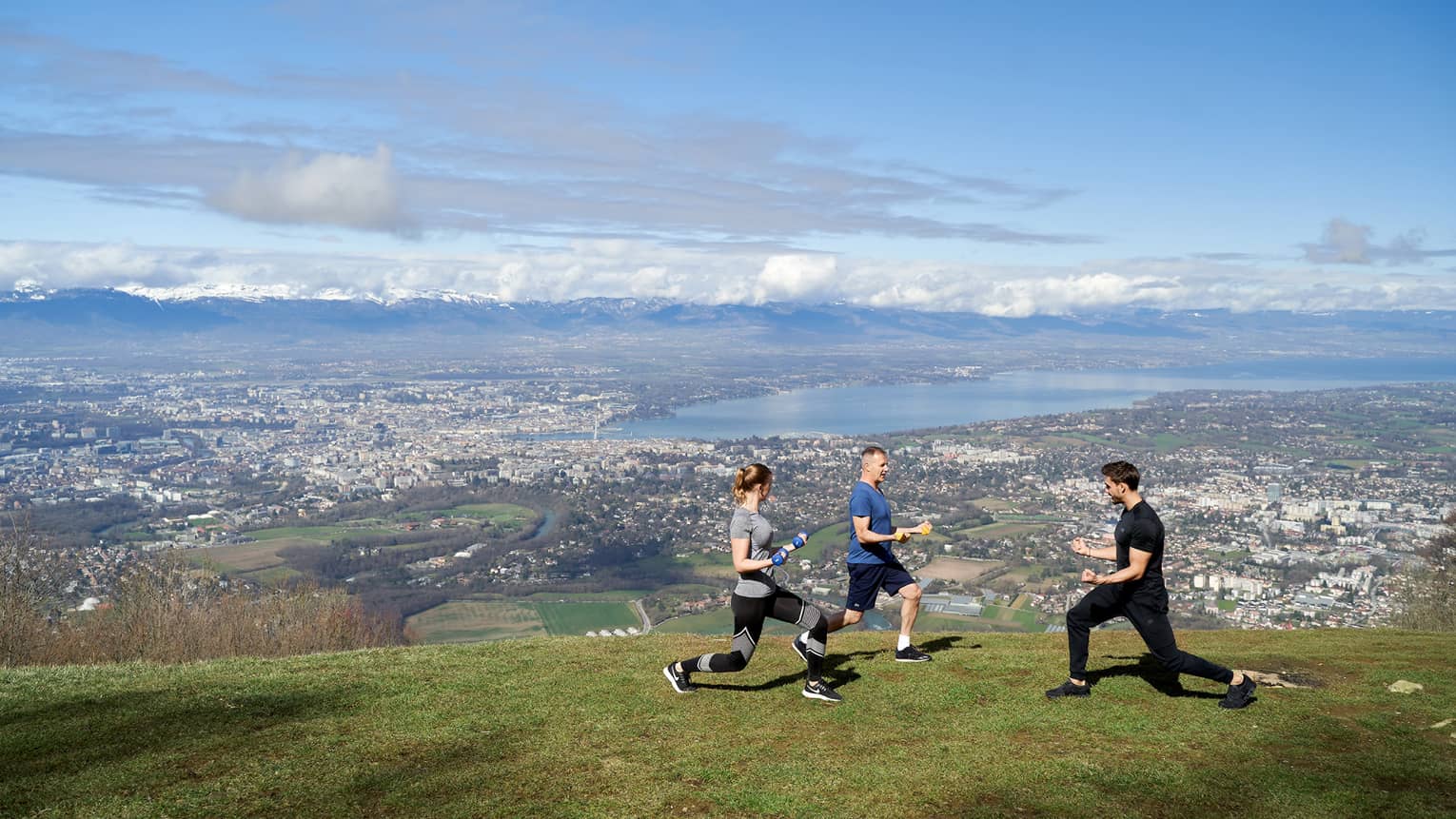 Two people on green mountainside doing knee bends with trainer and blue skies in background