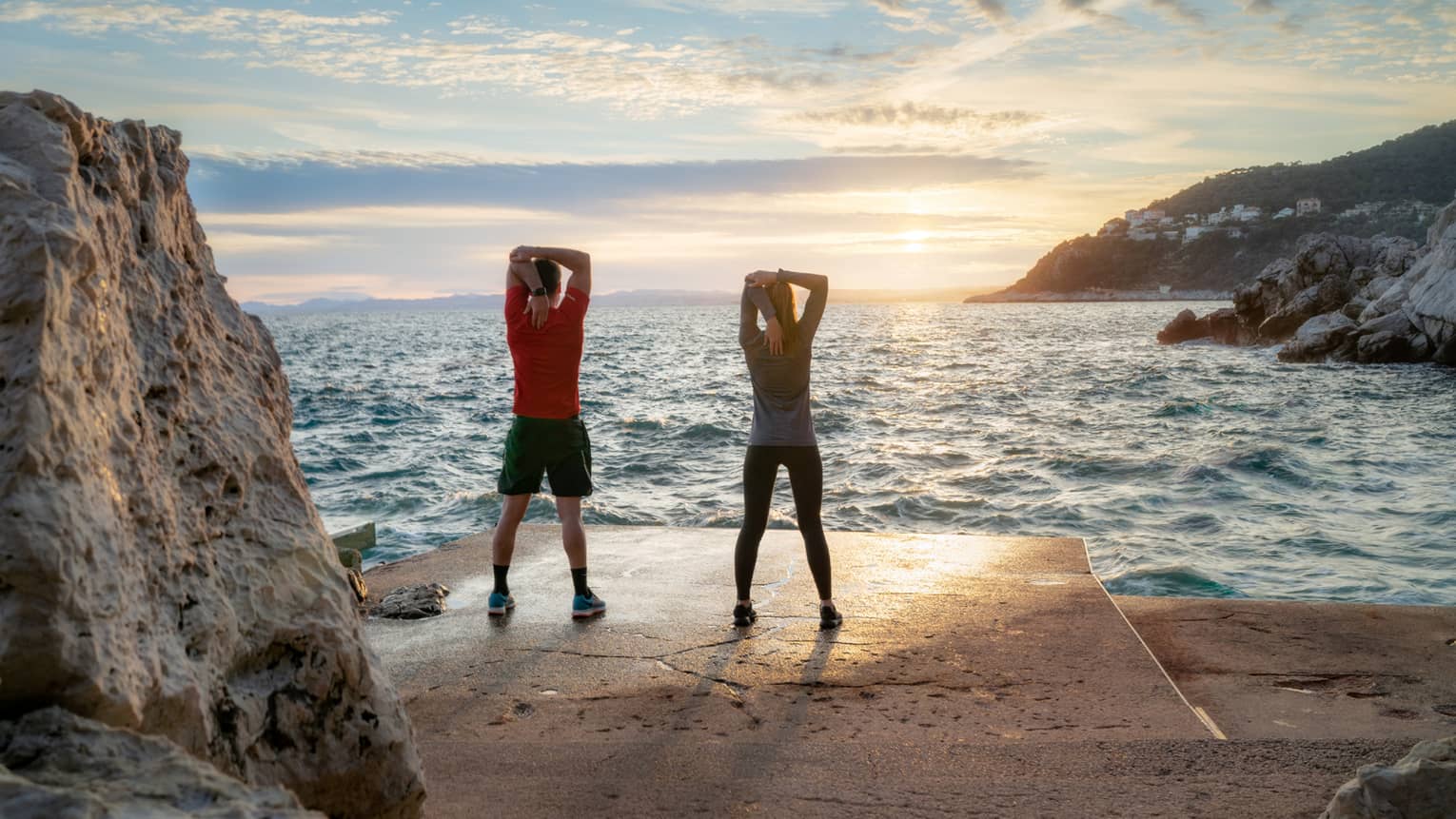 Rear view of two people standing and stretching at Mediterranean's edge at sunrise