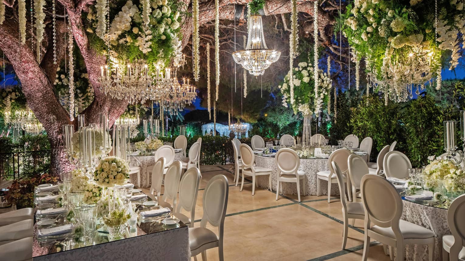 Four elegantly set mirrored-top tables with white chairs, crystal chandelier, floral arrangements hanging from ceiling