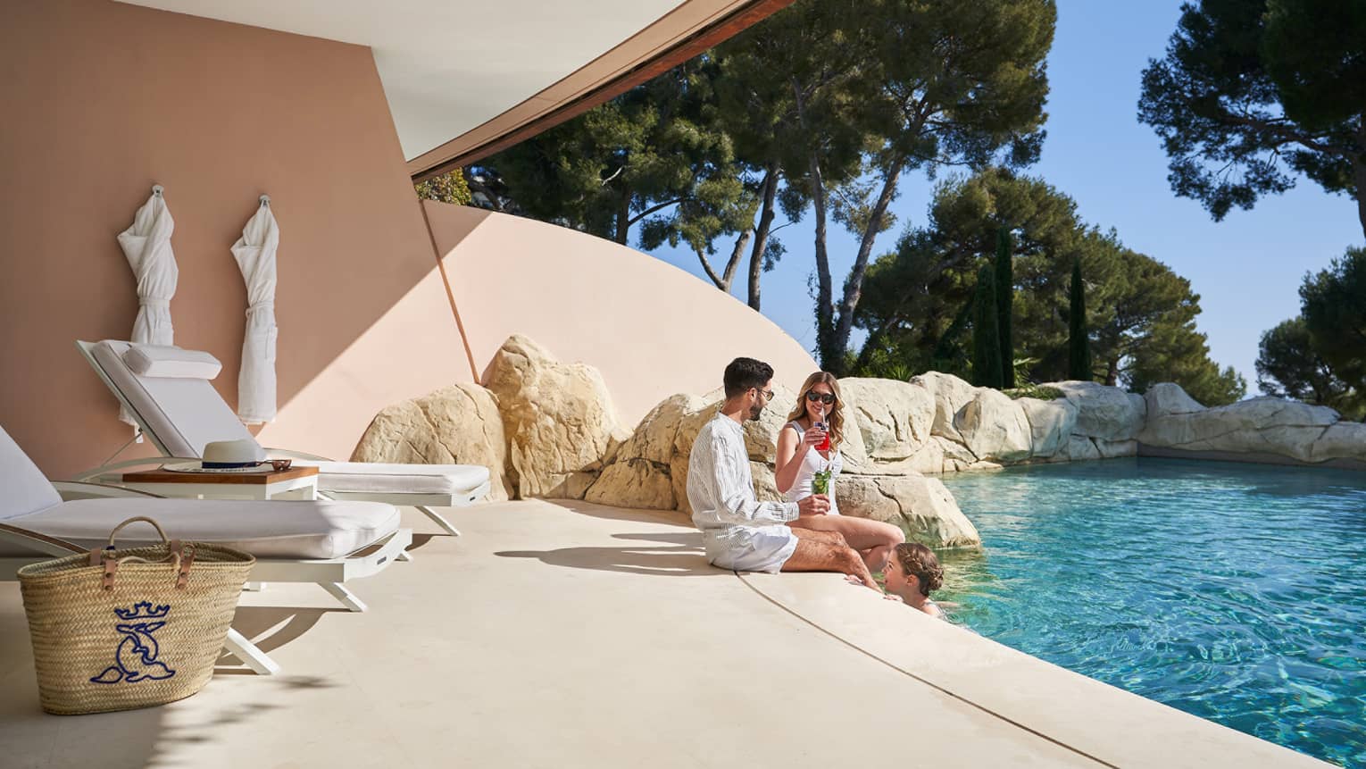 A family lounges on a terrace by an outdoor pool, surrounded by lush green trees
