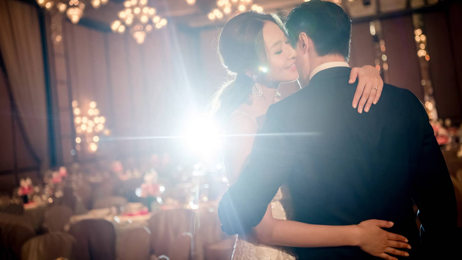 A bride and groom embrace each other in an indoor reception hall decorated with chandeliers and banquet tables