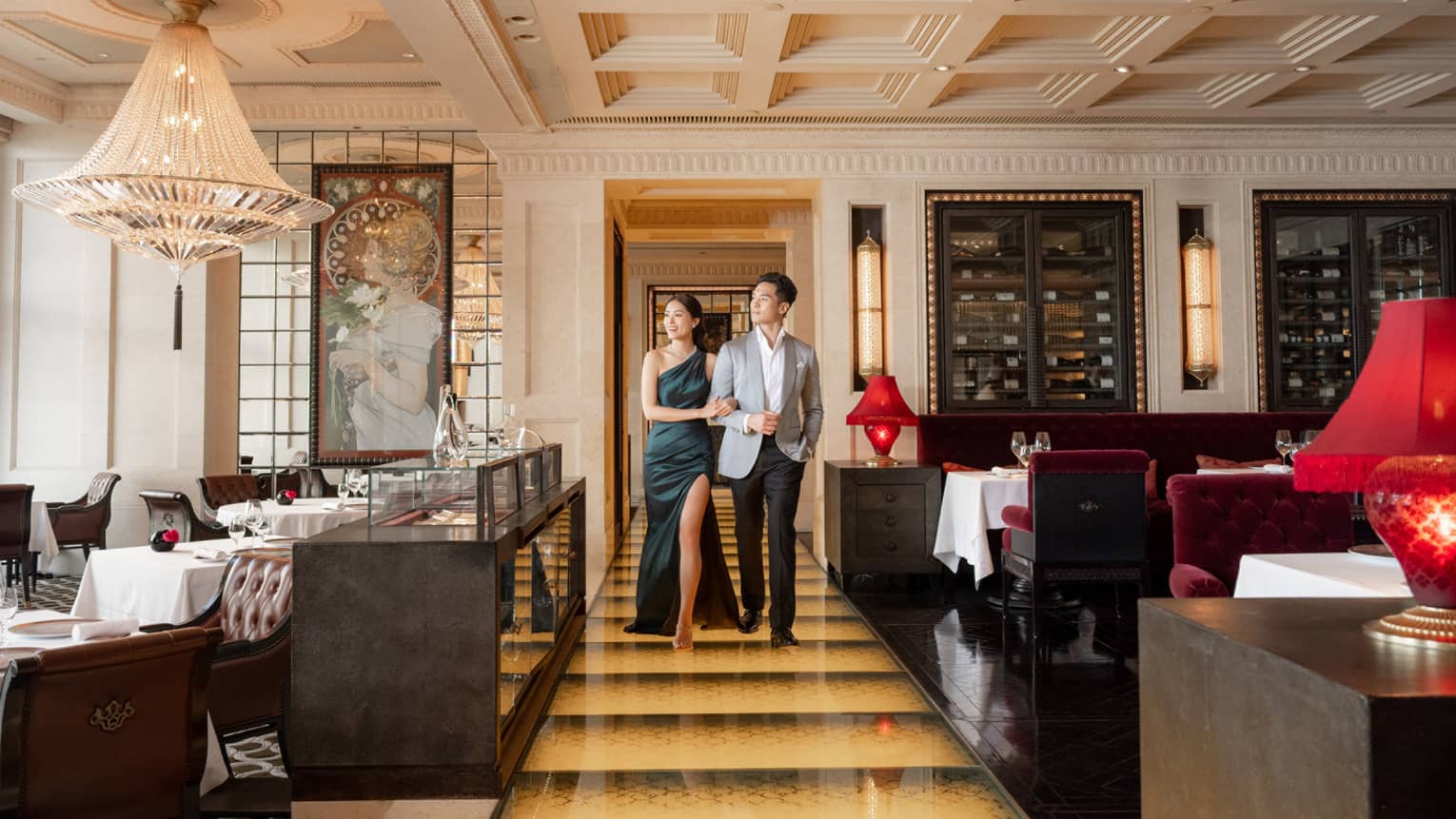 Elegant couple walking arm-in-arm in luxurious restaurant with chandeliers, red velvet seating and ornate decor.