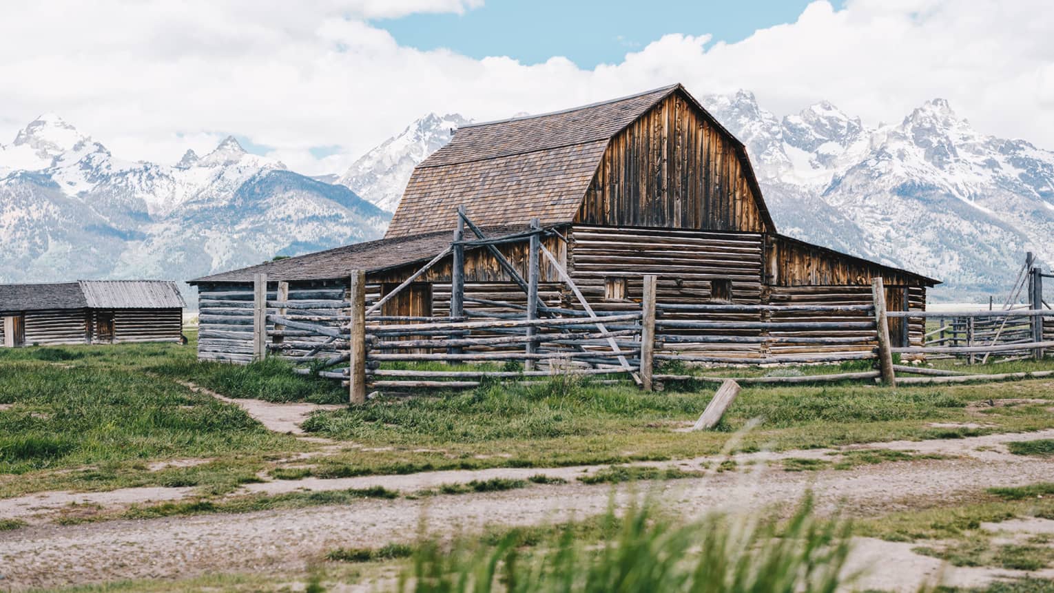 An old barn sits on a grassy patch of land, the snow-capped mountains looming behind it 