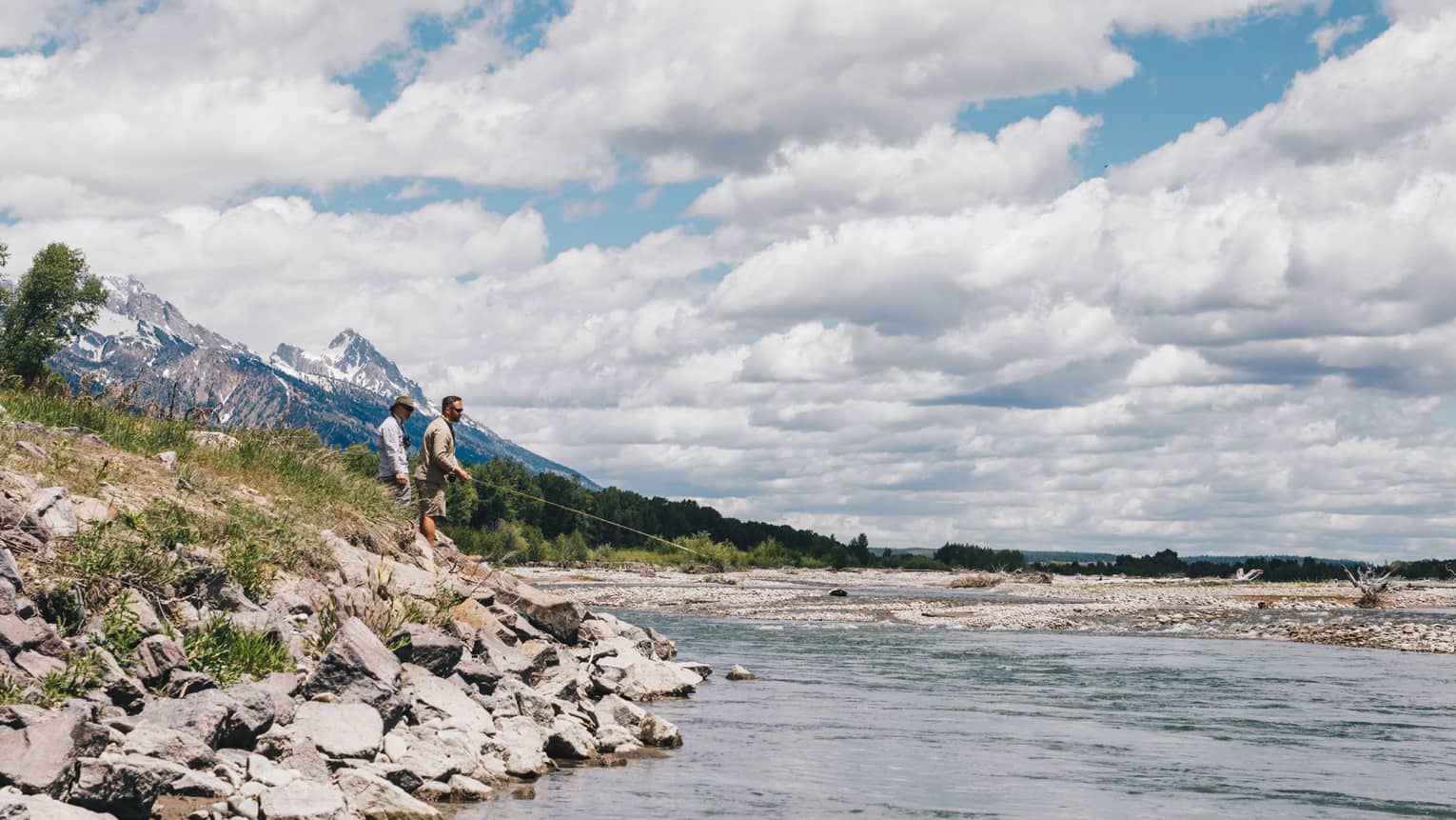 Two men fish on the rocky bank of a lake with fluffy white clouds above them in the sky