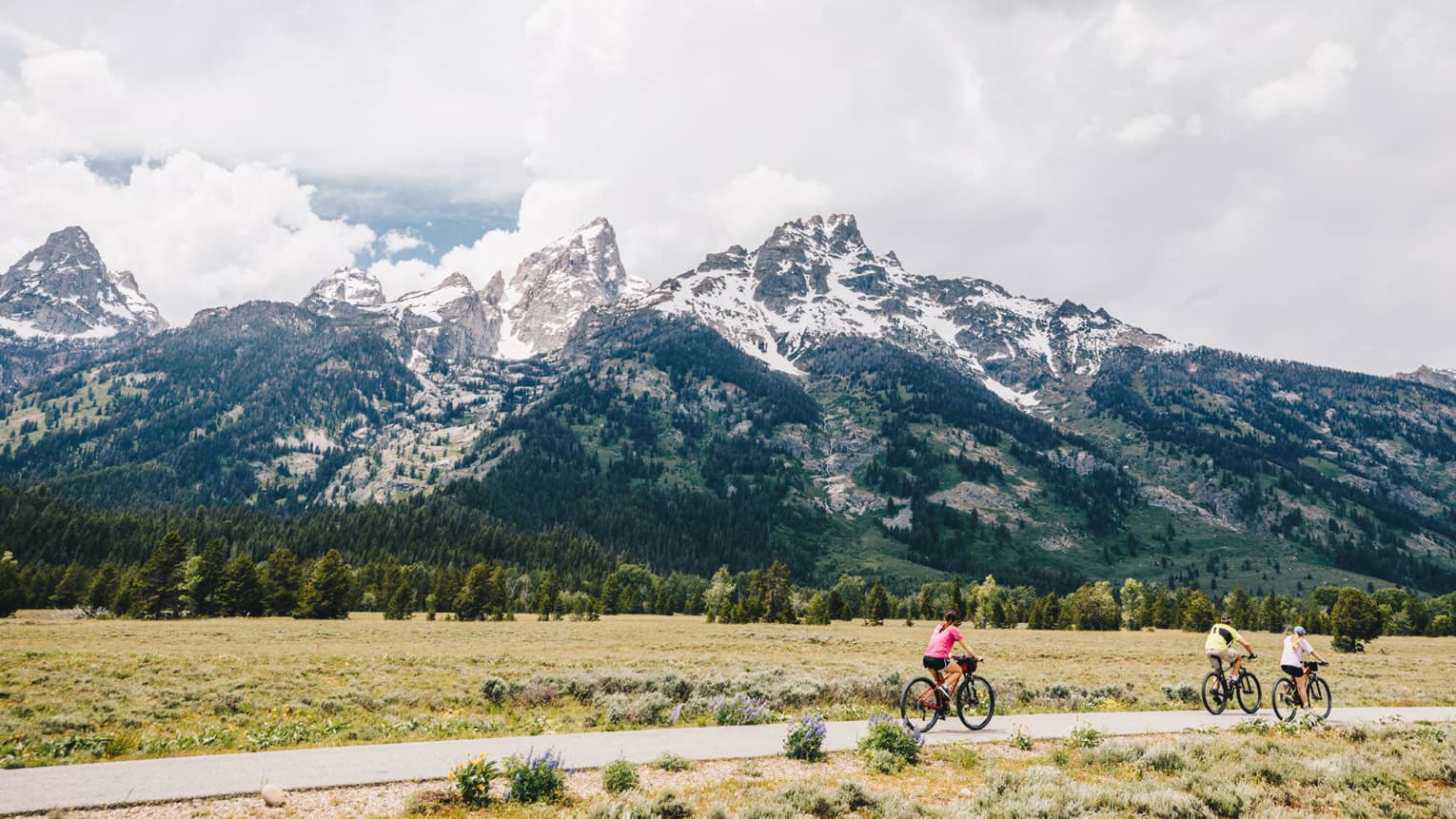 A man and two women cycle down a path on an overcast day with the mountains in the background 