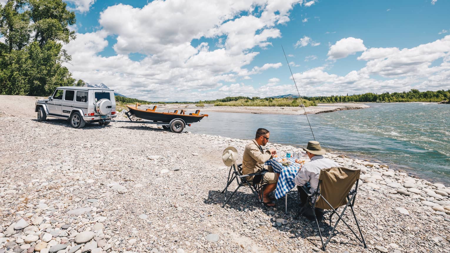 Two fishermen sit at a small table for a lakeside picnic, their boat hitched to their SUV in the background