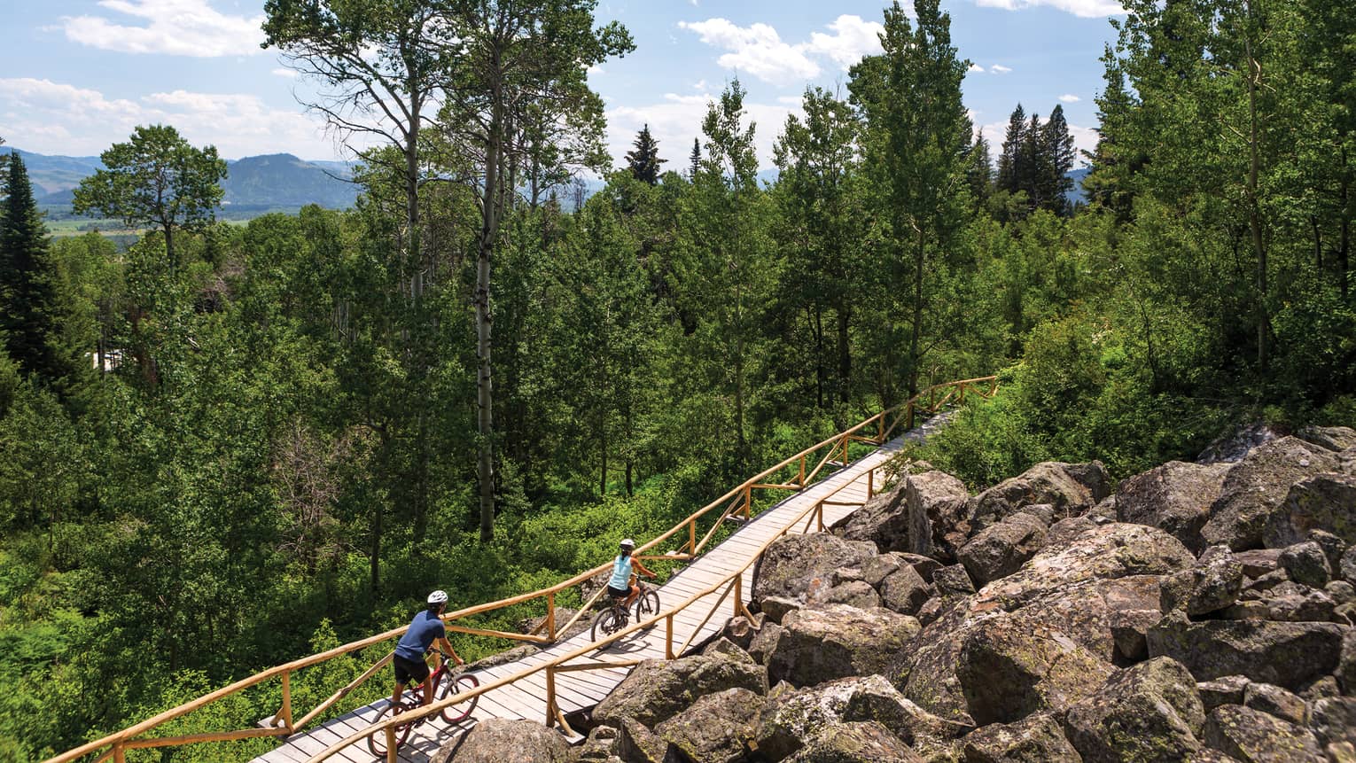 Aerial view of two cyclists on a forested bike path with mountains in the background