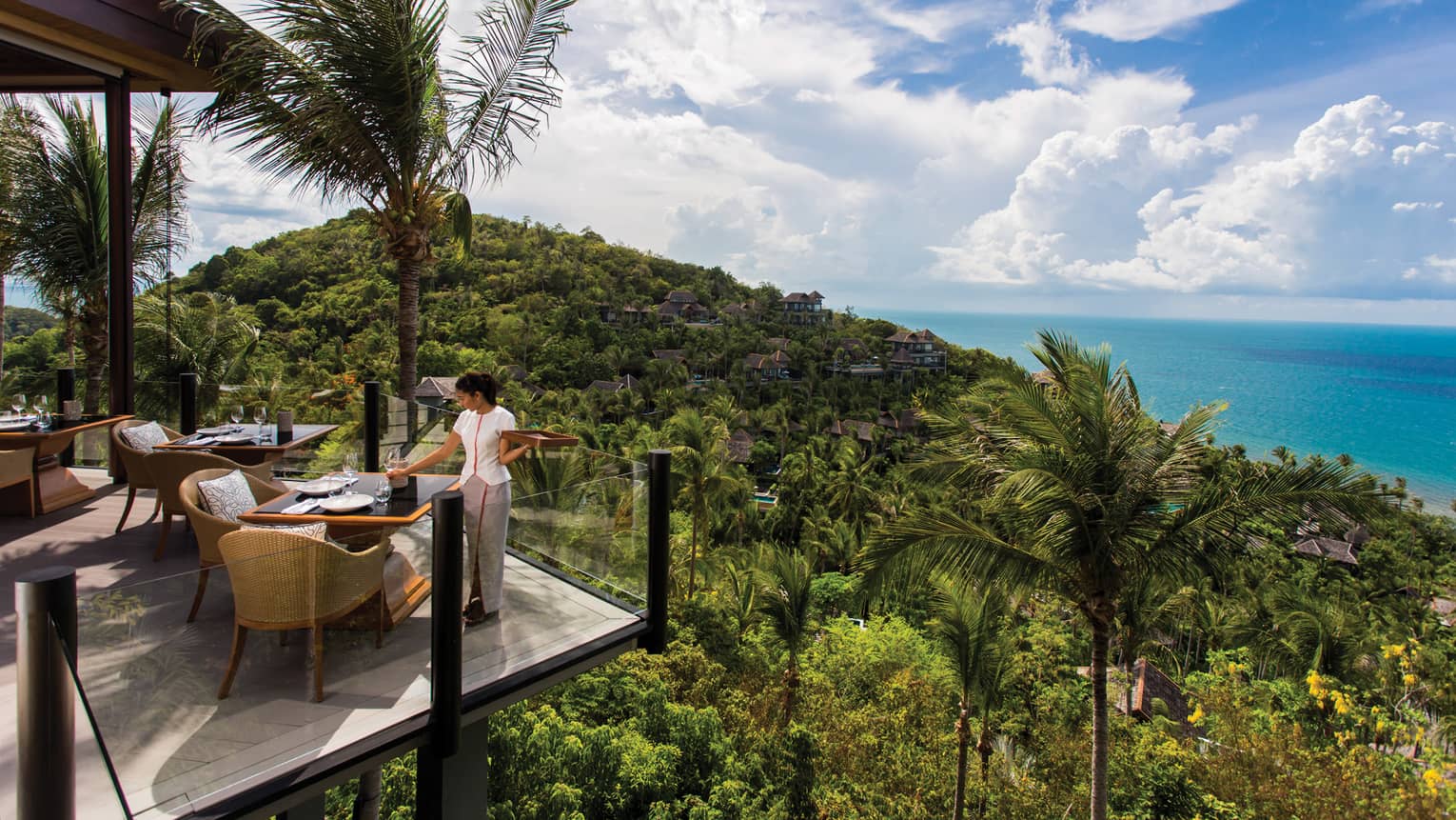 Server sets corner patio table behind glass balcony, high on tropical mountain