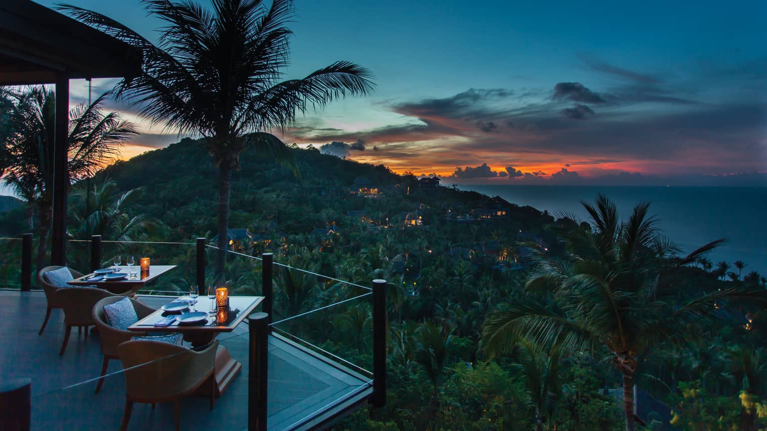 Corner of extended balcony with dining tables and chairs at dusk, expanse of forest, palm trees