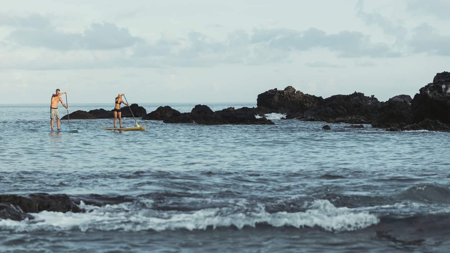 Two people balance on stand-up paddleboards in lagoon by black rocks