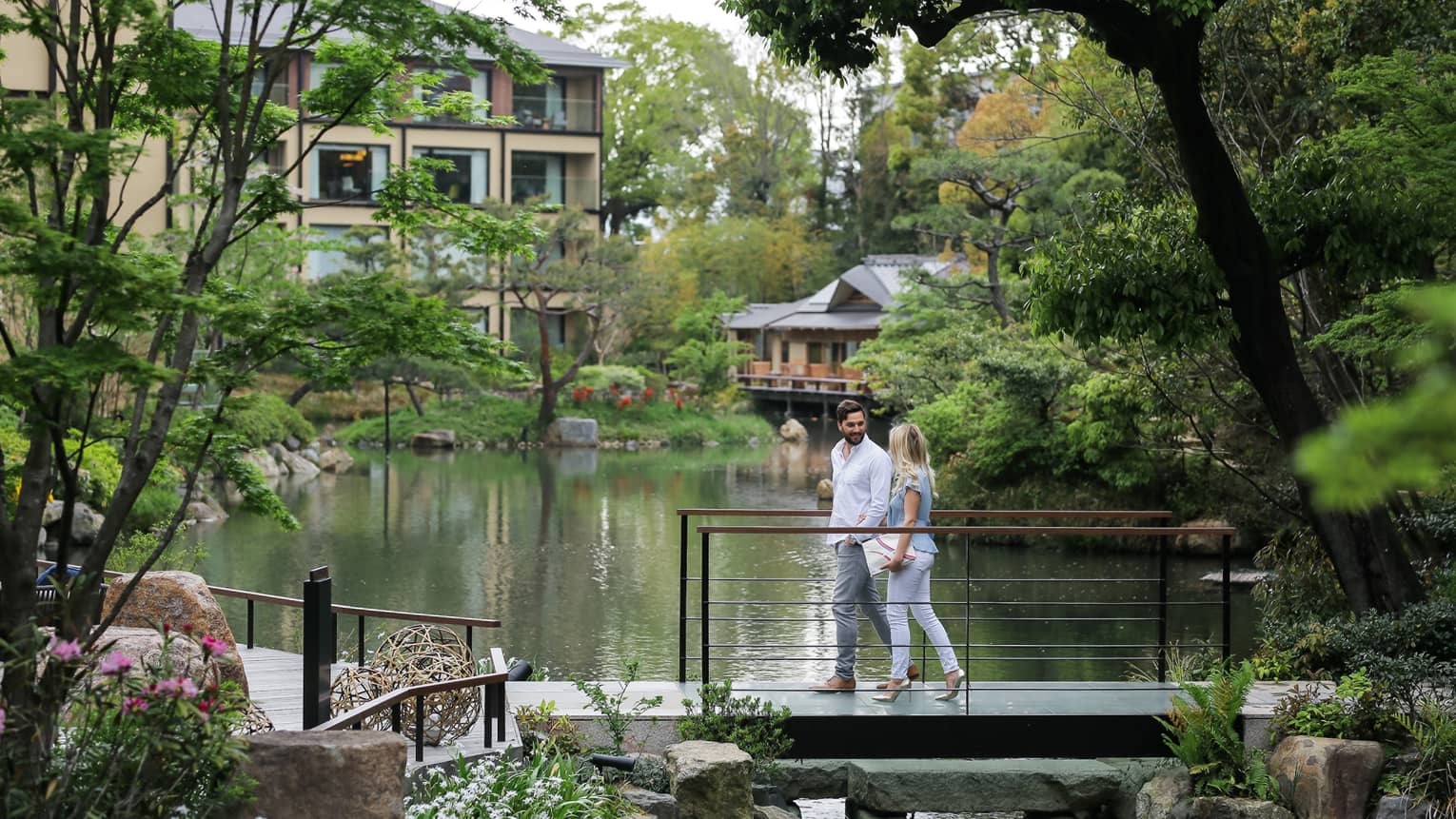 A couple leisurely walks across a footbridge, engaged in a conversation, overlooking a tranquil pond surrounded by lush greenery in a park.