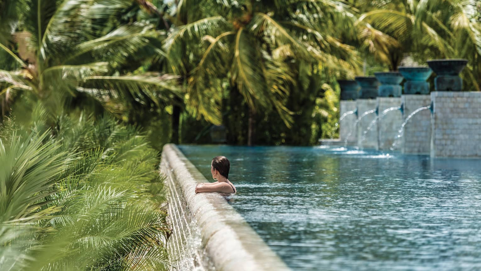 Woman wades, looks out from edge of outdoor swimming pool with fountains