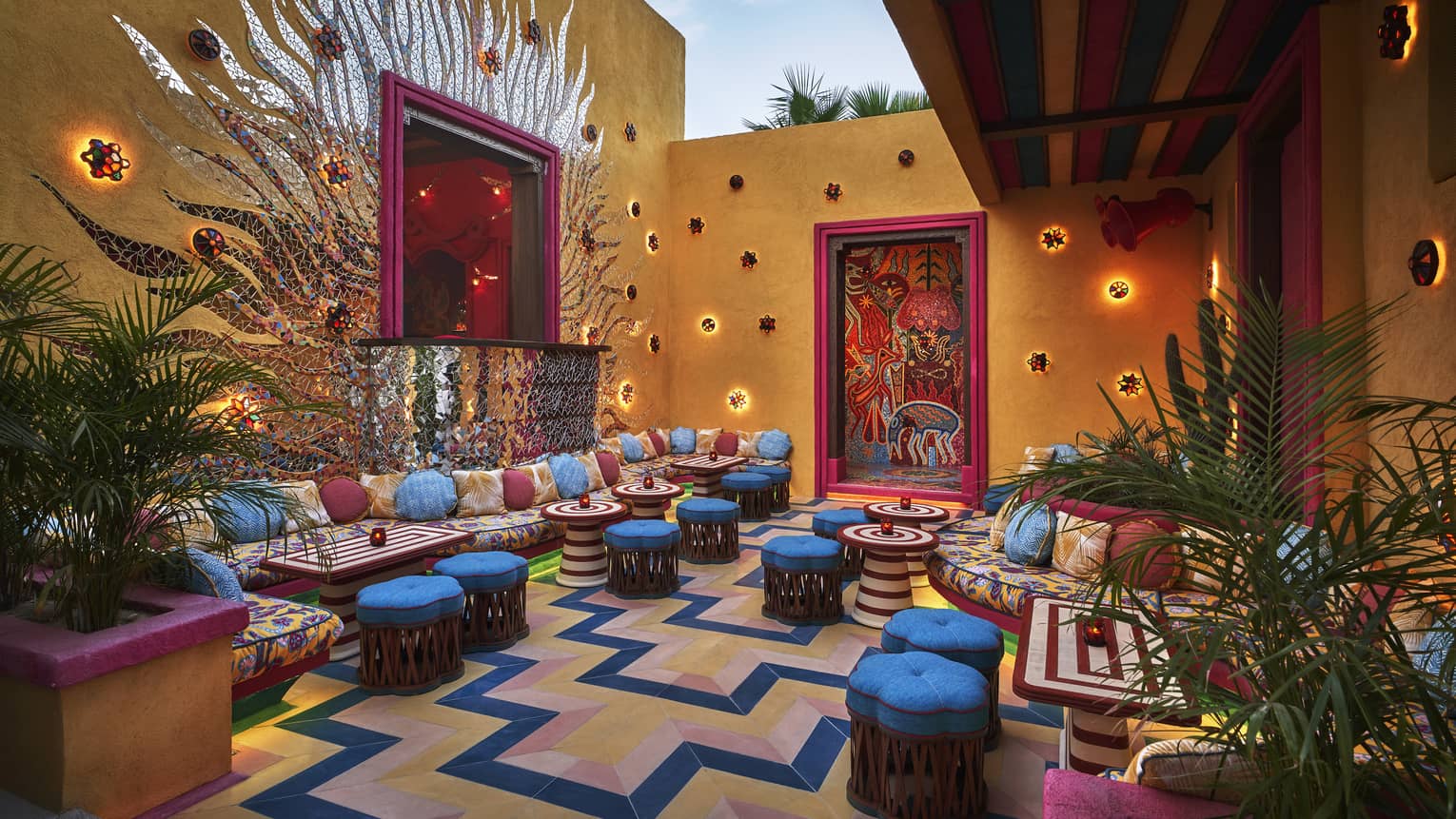 A colourful outdoor seating area with bright blue cushioned stools, colourful lights and plants.