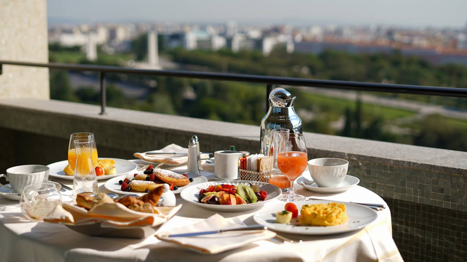 Outdoor breakfast table with pastries, fruit, juice and coffee, overlooking a city skyline and green parkland.