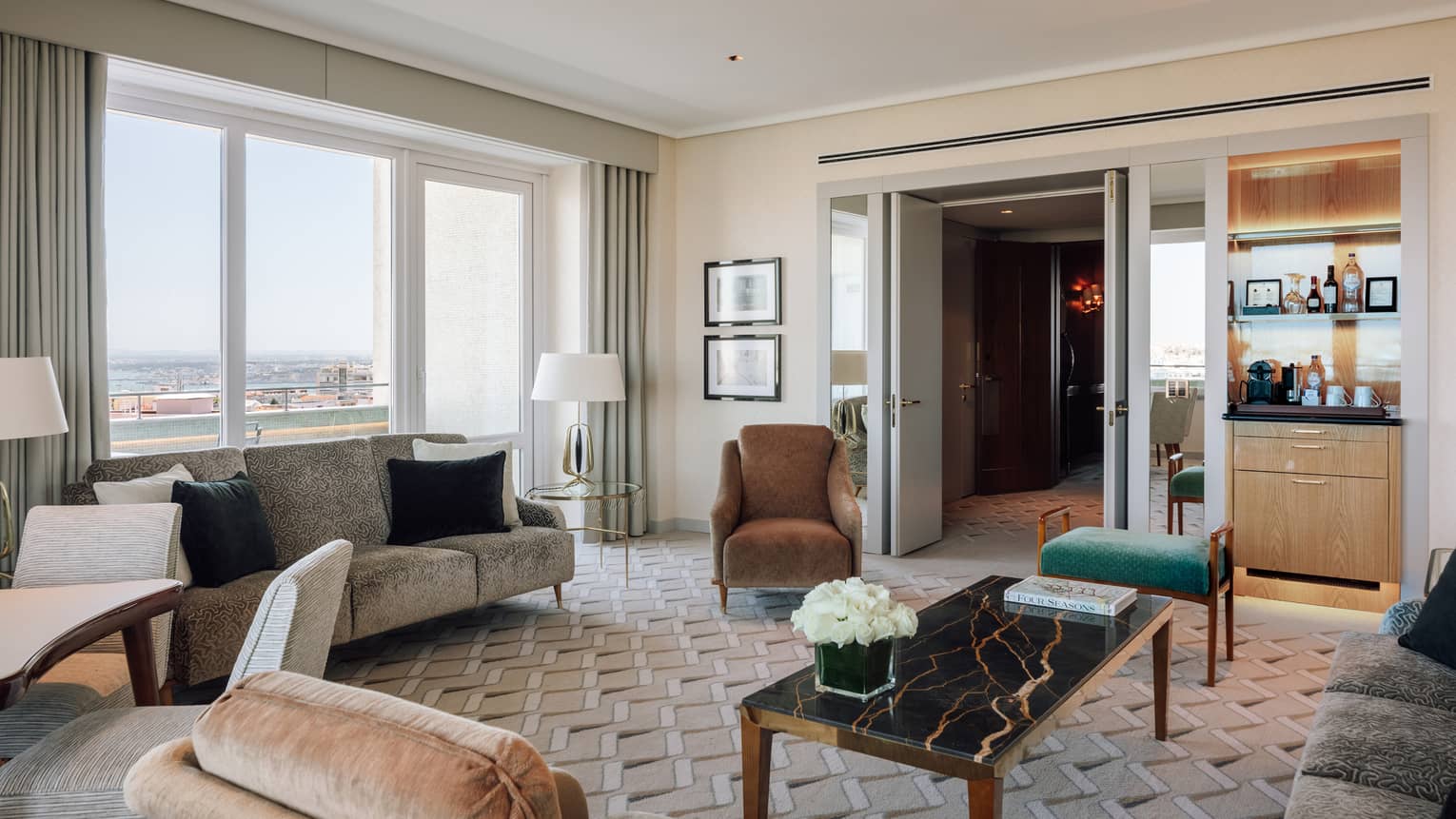 Elegant hotel suite living room with sofas, armchairs, a coffee table, minibar and large windows with city views.