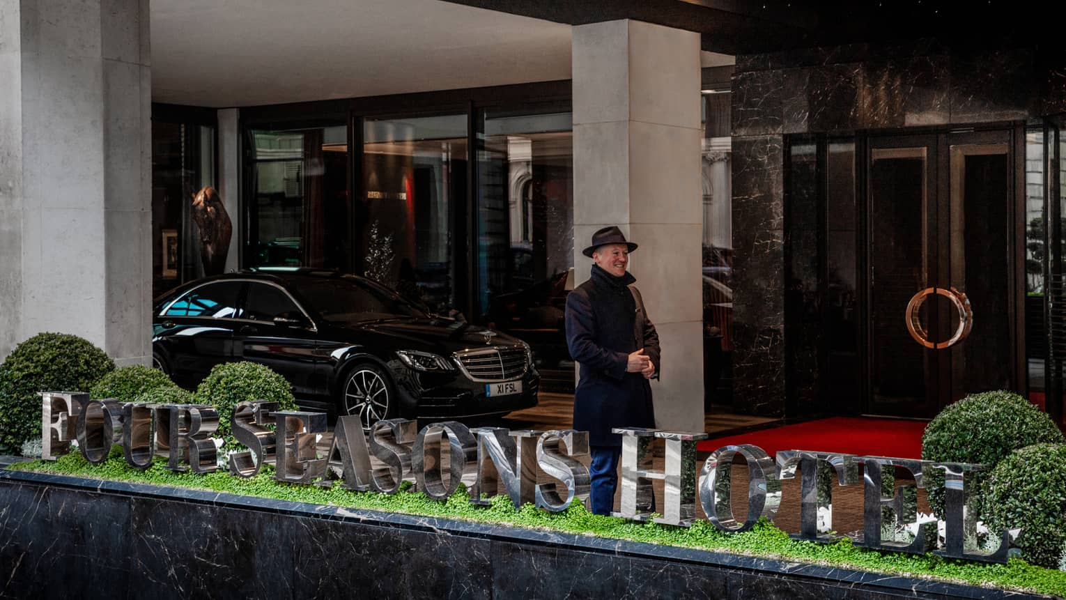 Man in dark winter coat and fedora, standing and smiling at hotel exterior under breezeway near black sedan and behind hotel sign