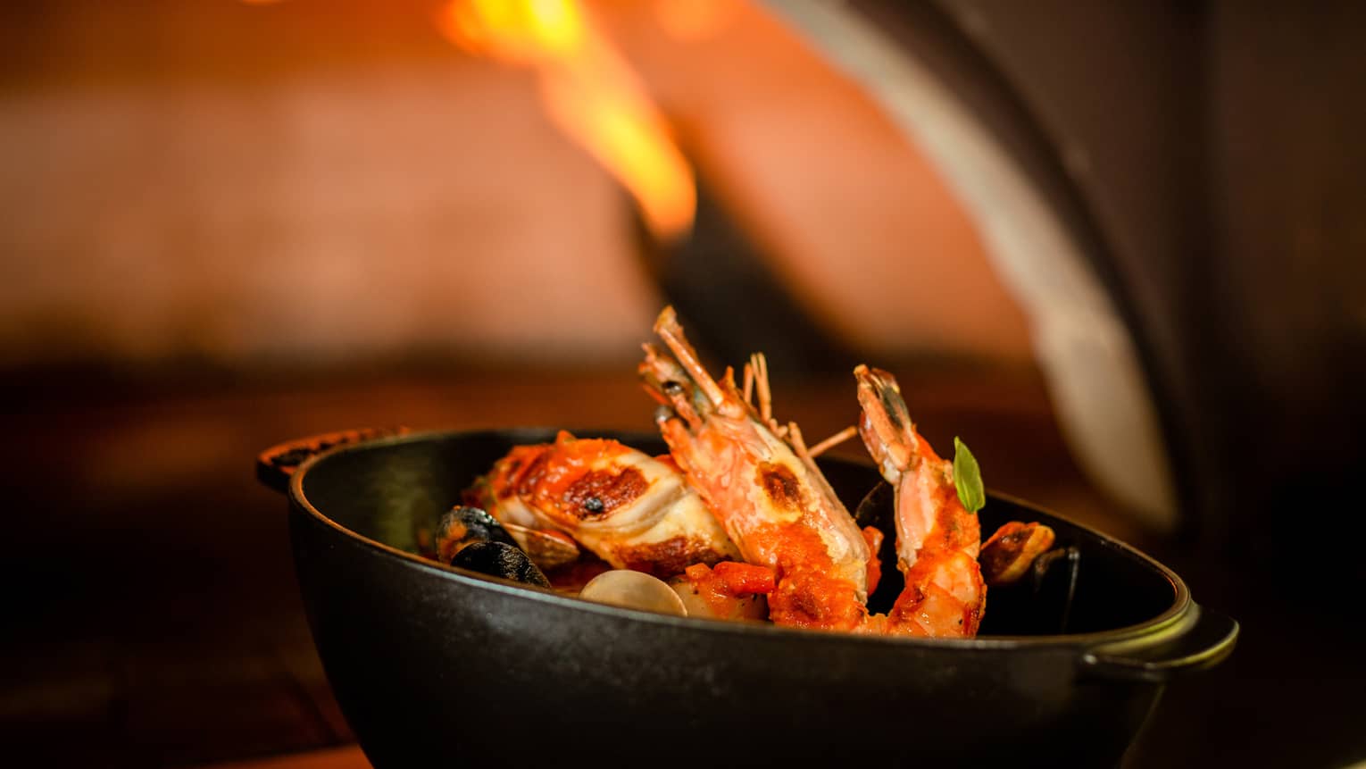 Cioppino with head-on prawns in cast iron serving dish in front of pizza oven