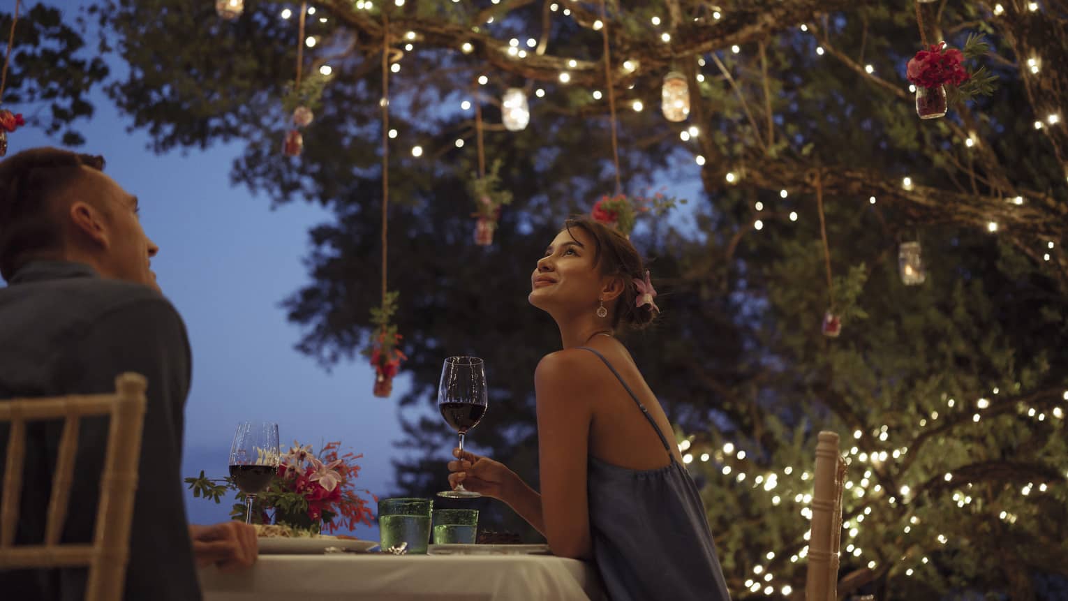 A smiling couple having a romantic dinner under trees covered in string lights and jars of flowers hanging from the branches.