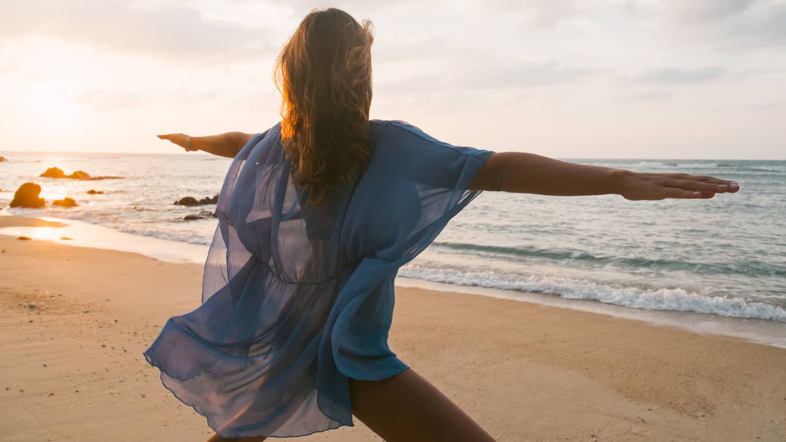 Woman wearing sheer beach cover kneels with arms outstretched in yoga pose on sandy beach by water