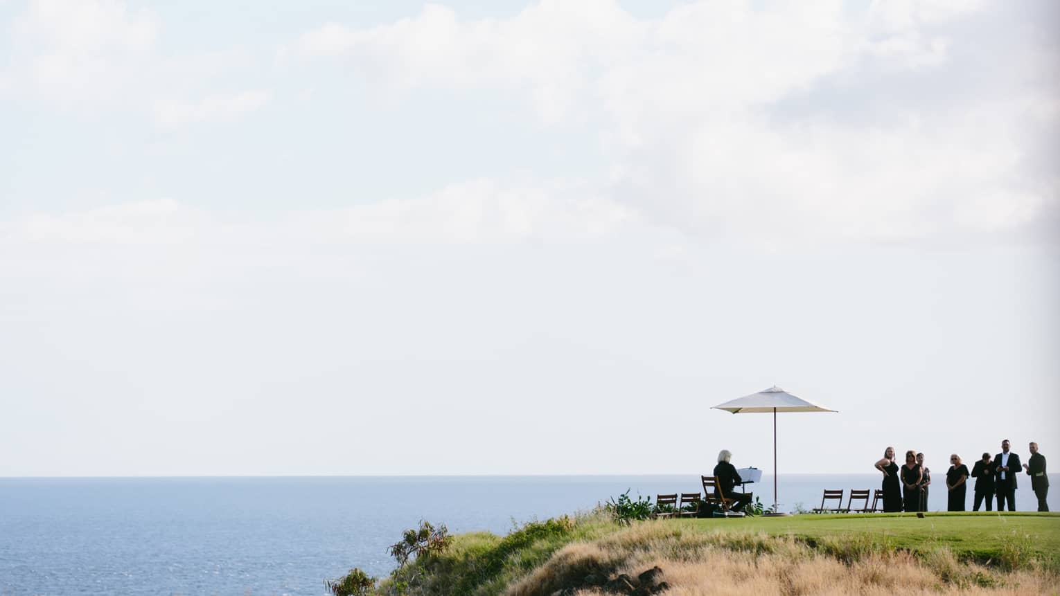 A group of people dressed in black congregate on the edge of a cliff overlooking a calm sea.