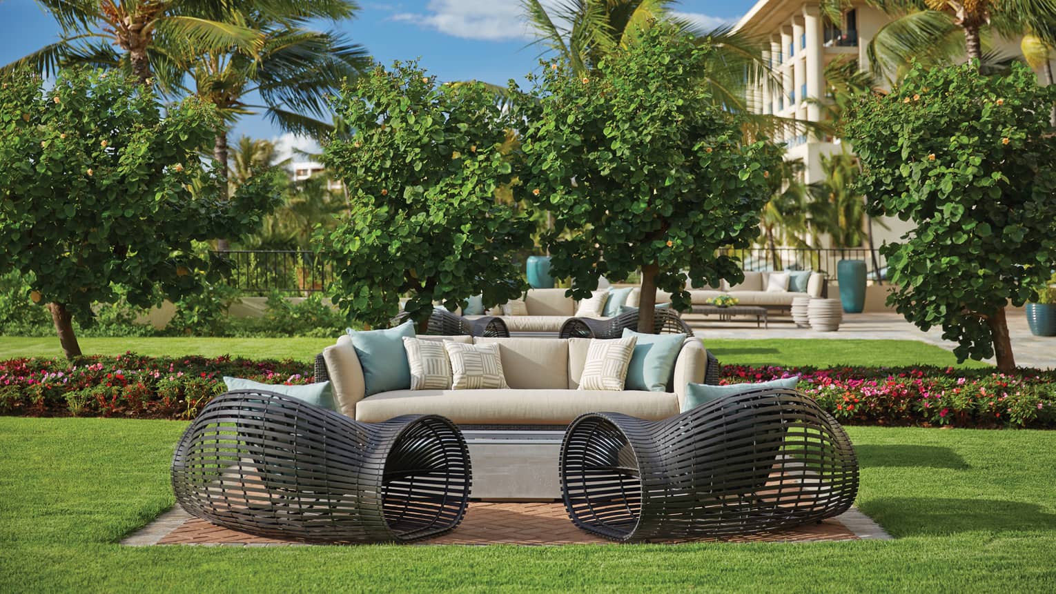 Outdoor seating area with cushioned sofa and modern black chairs on lawn surrounded by trees and flowers