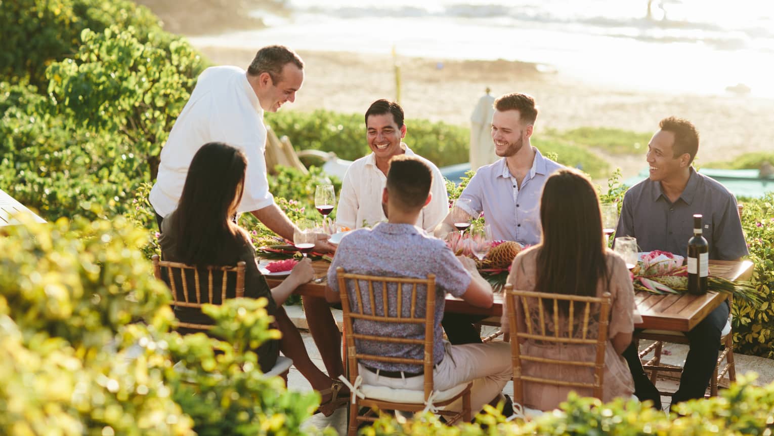 Six people smiling, sit around outdoor wooden table by ocean as wait staff serves them