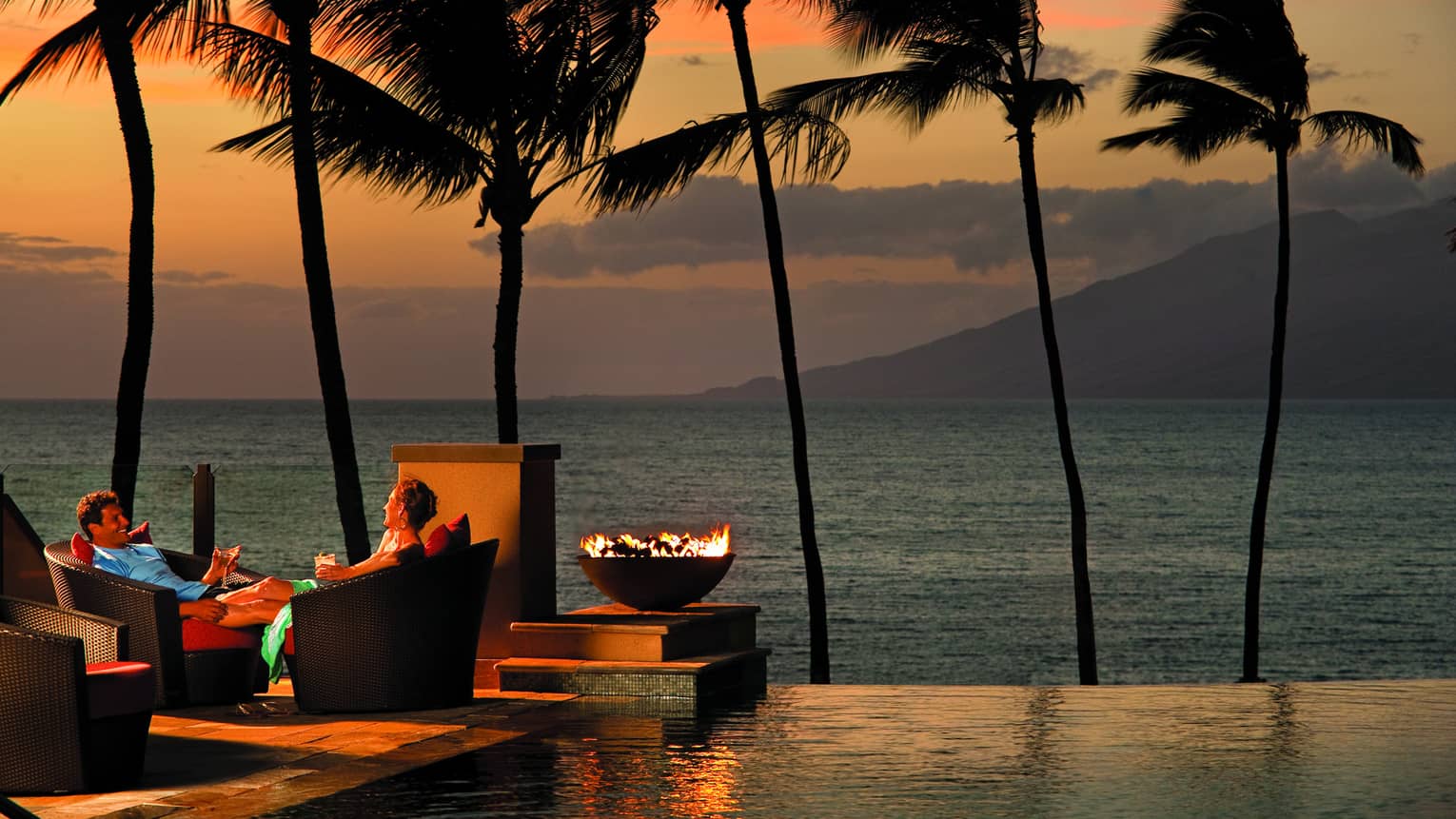 Man and woman sit in wicker patio chairs with cocktails at sunset, next to infinity pool and fire pit