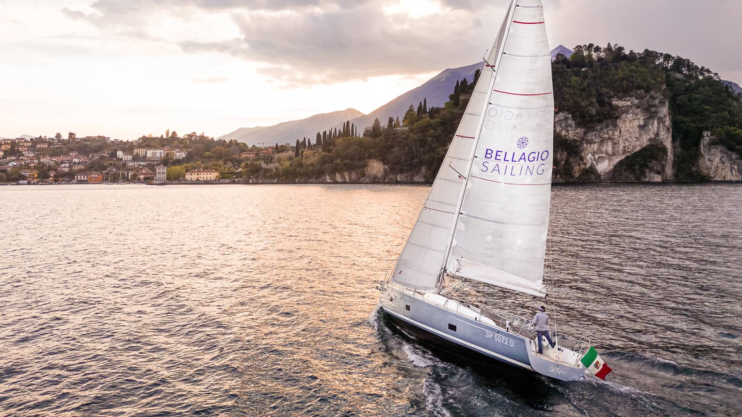 A sailboat on water near region with trees and buildings.