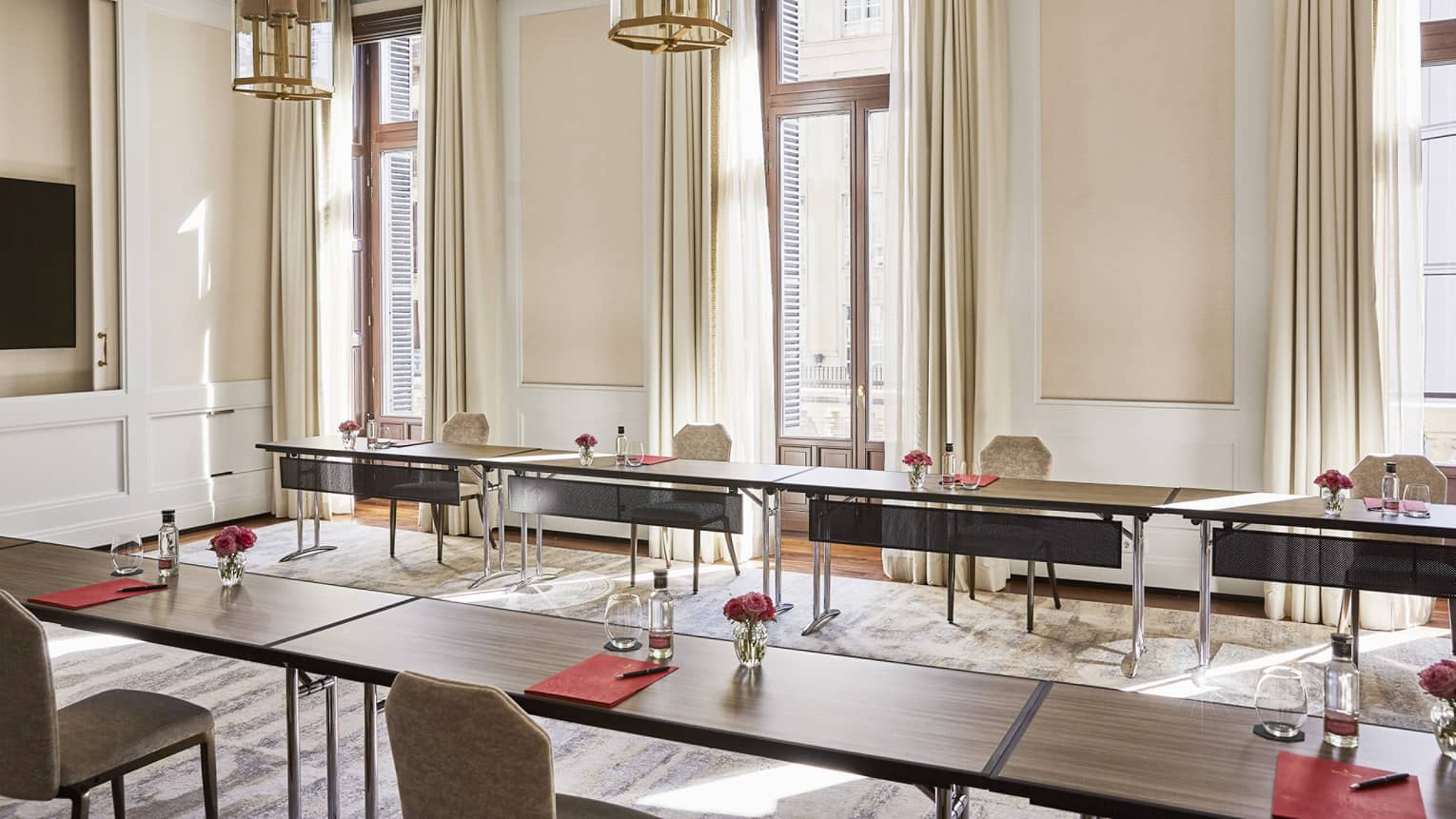 A meeting room with a flat-screen TV on one wall and large windows admitting natural light on another wall is decorated in warm neutral colors and has chandelier lighting, chairs, and narrow brown tables set with red folders, pens, drinking glasses, clear bottles, and red flowers in clear vases