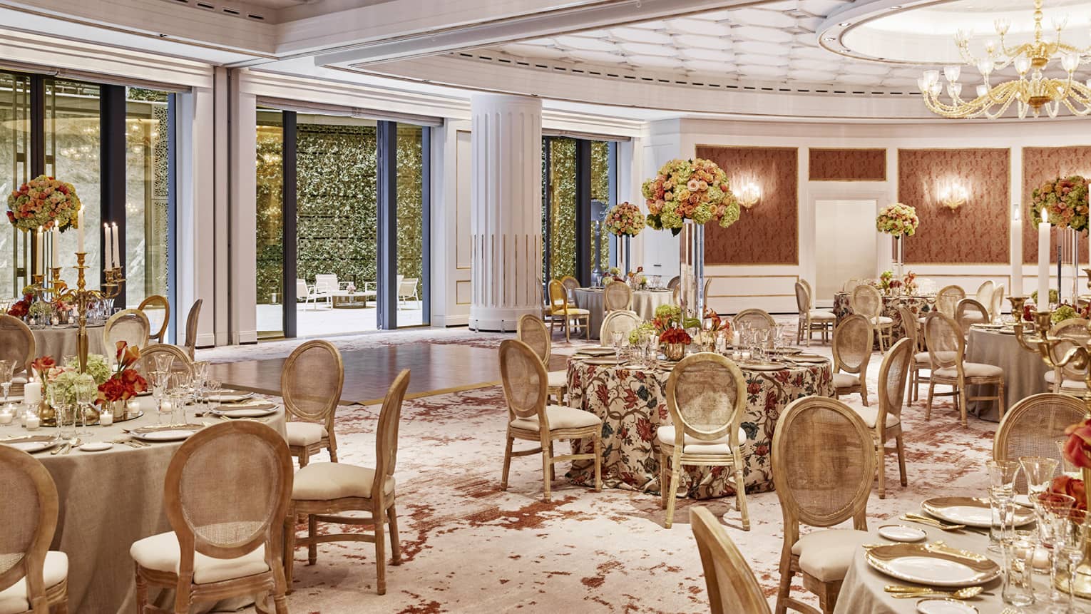 A luxurious banquet hall set for a formal event, featuring round tables with floral centrepieces, elegant chairs, chandeliers and large windows overlooking a garden.