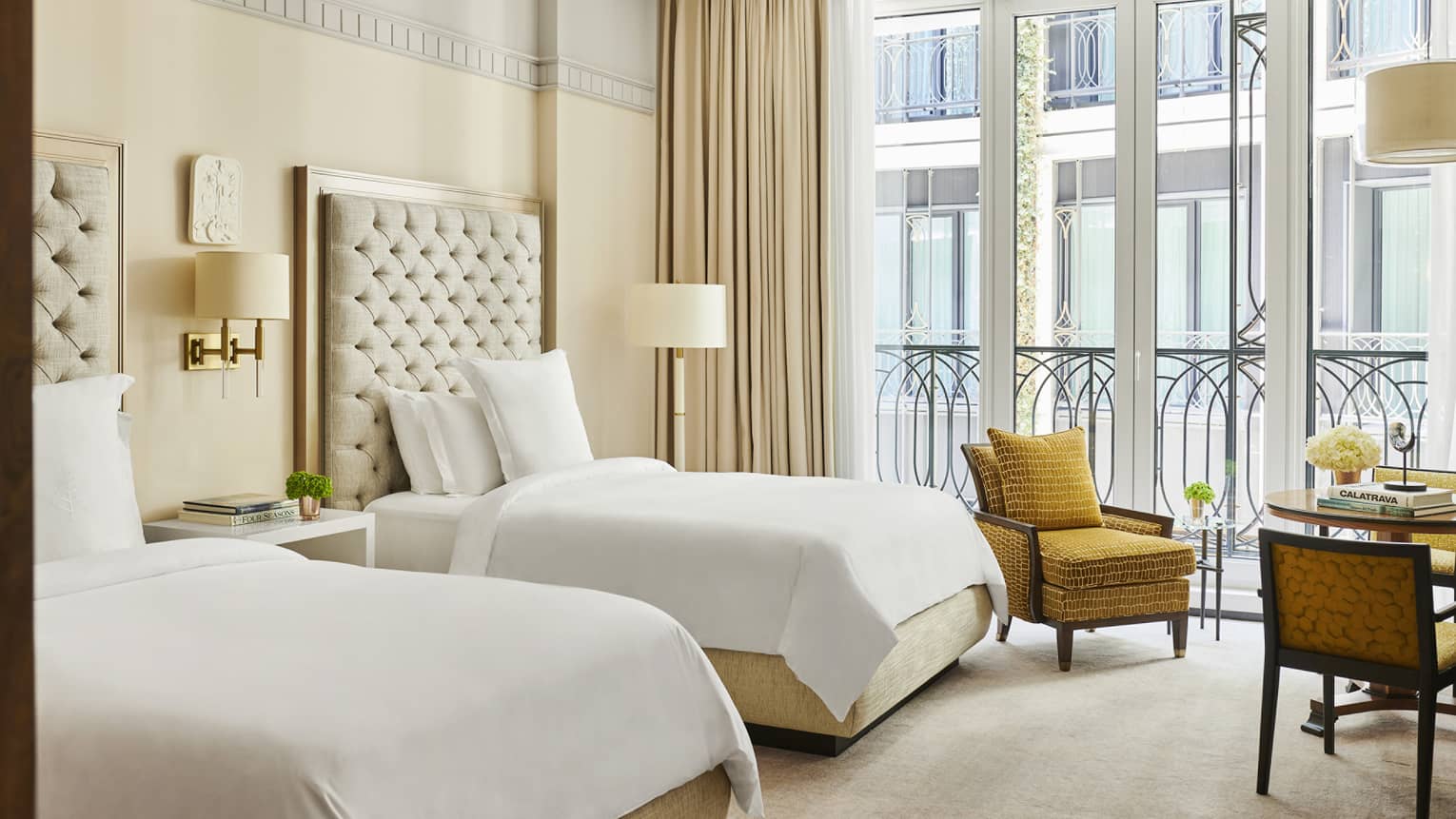 Elegant hotel room with two beds, tufted headboards, a mustard armchair, small round table and large windows with decorative railing and beige curtains