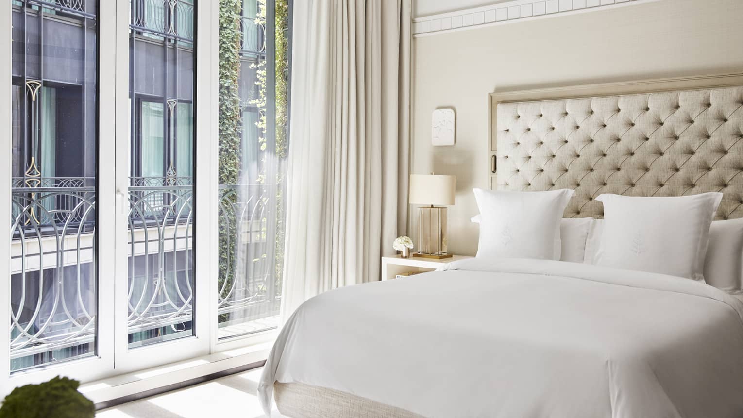 Luxury hotel bedroom with a king bed, tufted headboard, bedside table with lamp and flowers and floor-to-ceiling windows with decorative railing and curtains