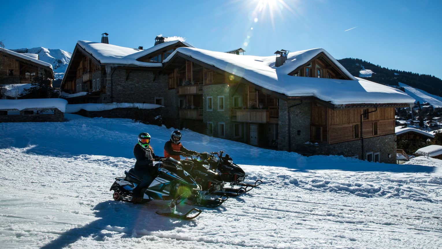 Pair of snowmobiles riding along snowy mountain trail past lodges on sunny day