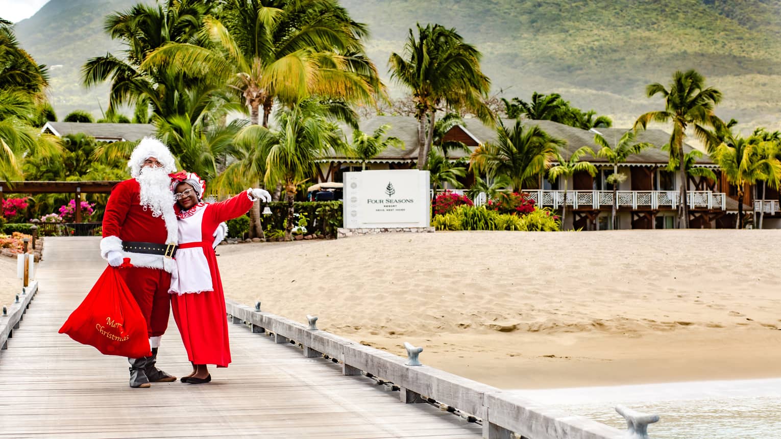 Couple dressed up as Santa and Mrs. Claus stand in front of Resort sign, mountains and palm trees visible in background
