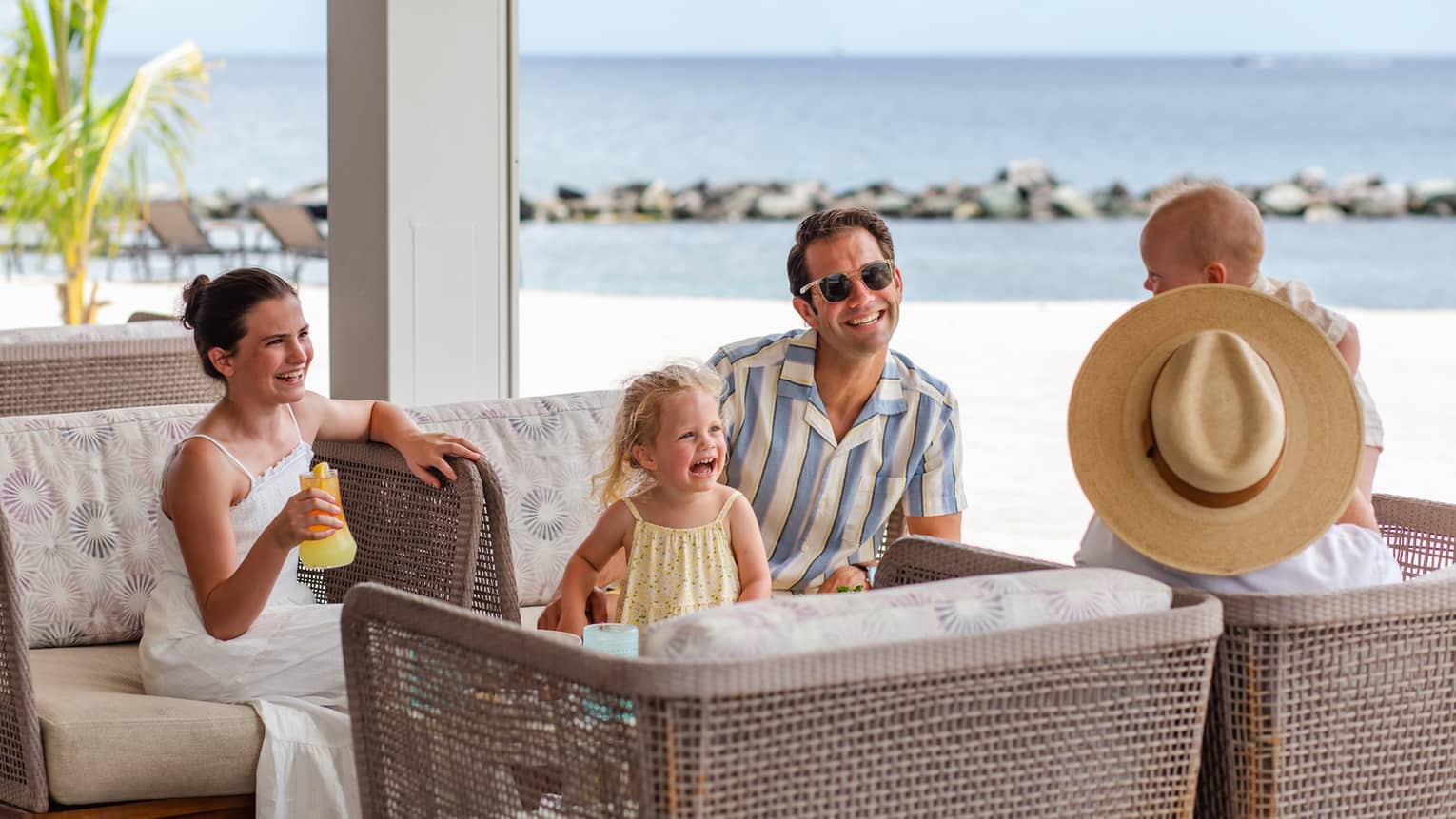 Three adults and two young children sit together facing each other on four rattan arm chairs with a beach in the background
