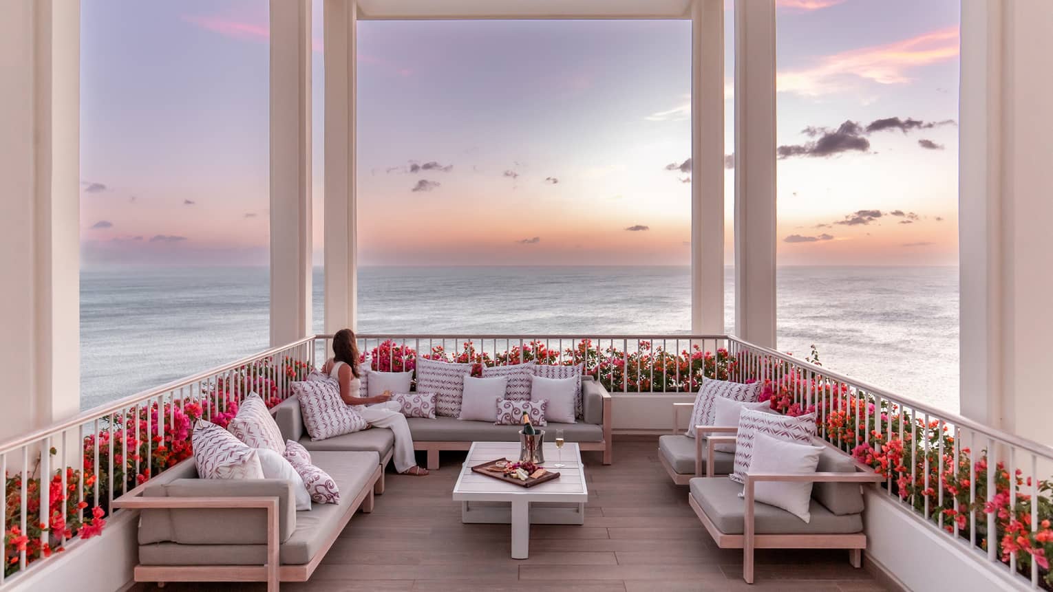 Woman sits on sofa on outdoor terrace under a white pergola overlooking the ocean