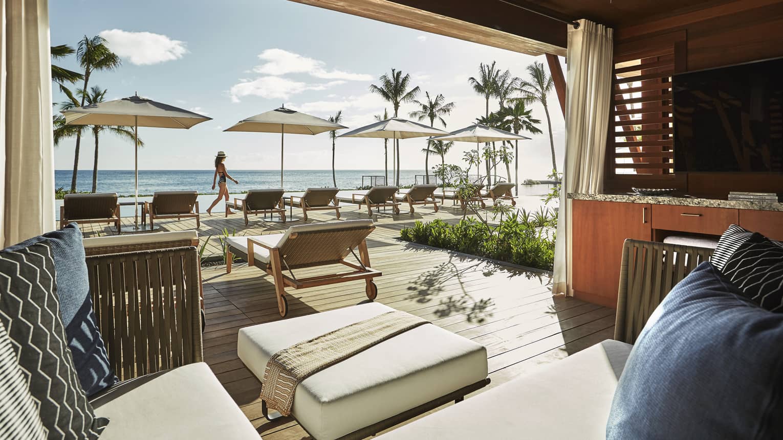 Plush patio lounge chairs under wood cabana, woman walks by pool deck