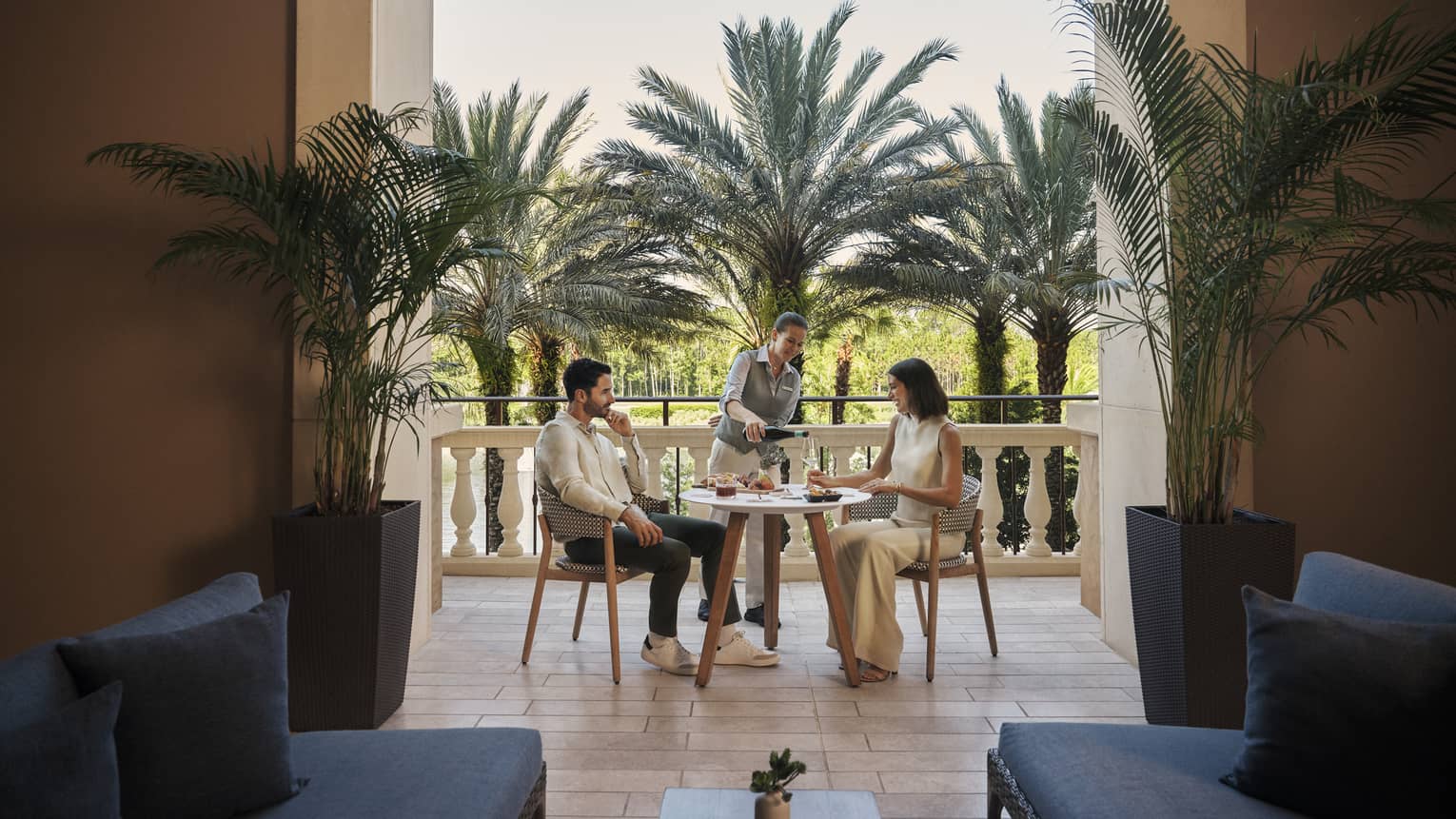 Two people sit at a small table surrounded by palm trees on a tiled patio near a large doorway