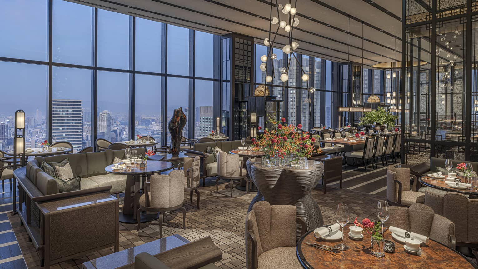 Expansive high-end restaurant warmly lit by modern chandeliers with a bank of windows offering aerial views of the city