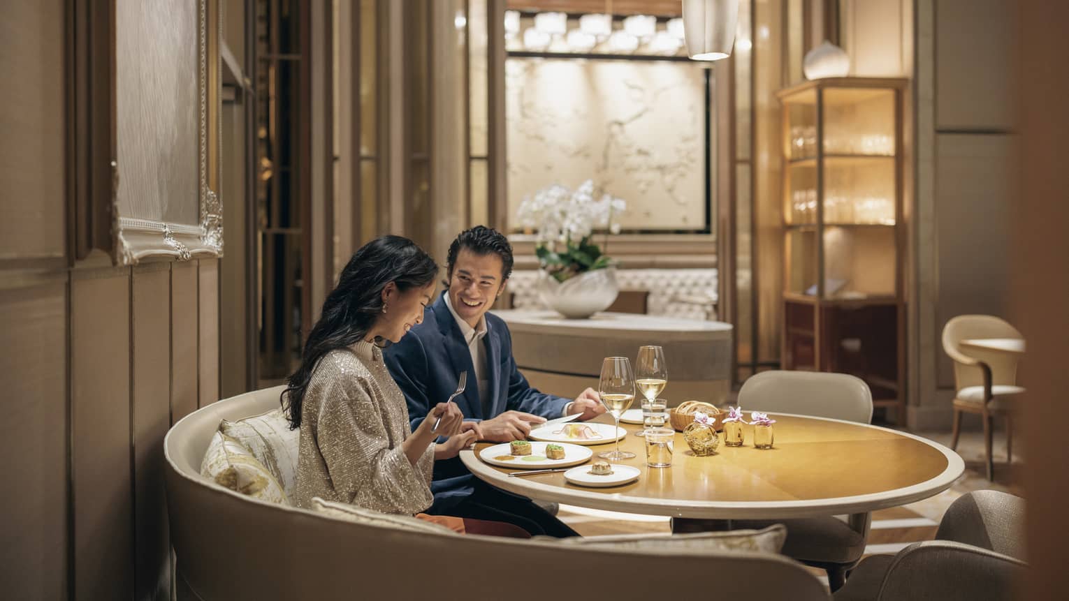A smiling couple savours lunch with wine in a circular booth at a fine dining restaurant with warm pendant lighting.