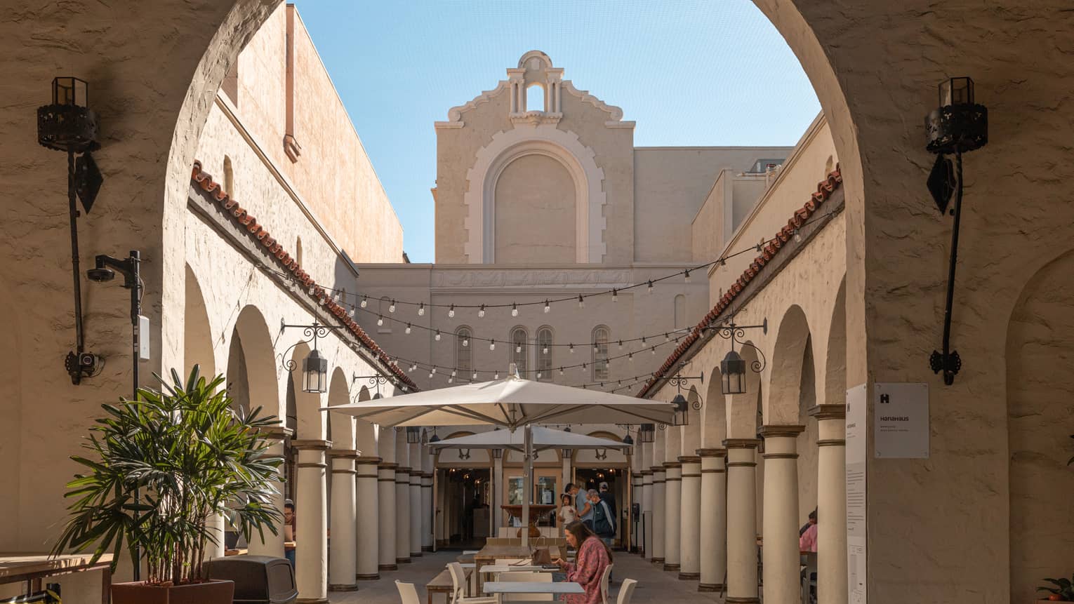 An archway leading to a small street with columns and surrounded by small tan buildings.
