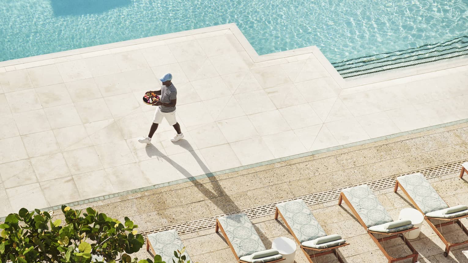 A team member walking next to a pool holding a tray of drinks and food.