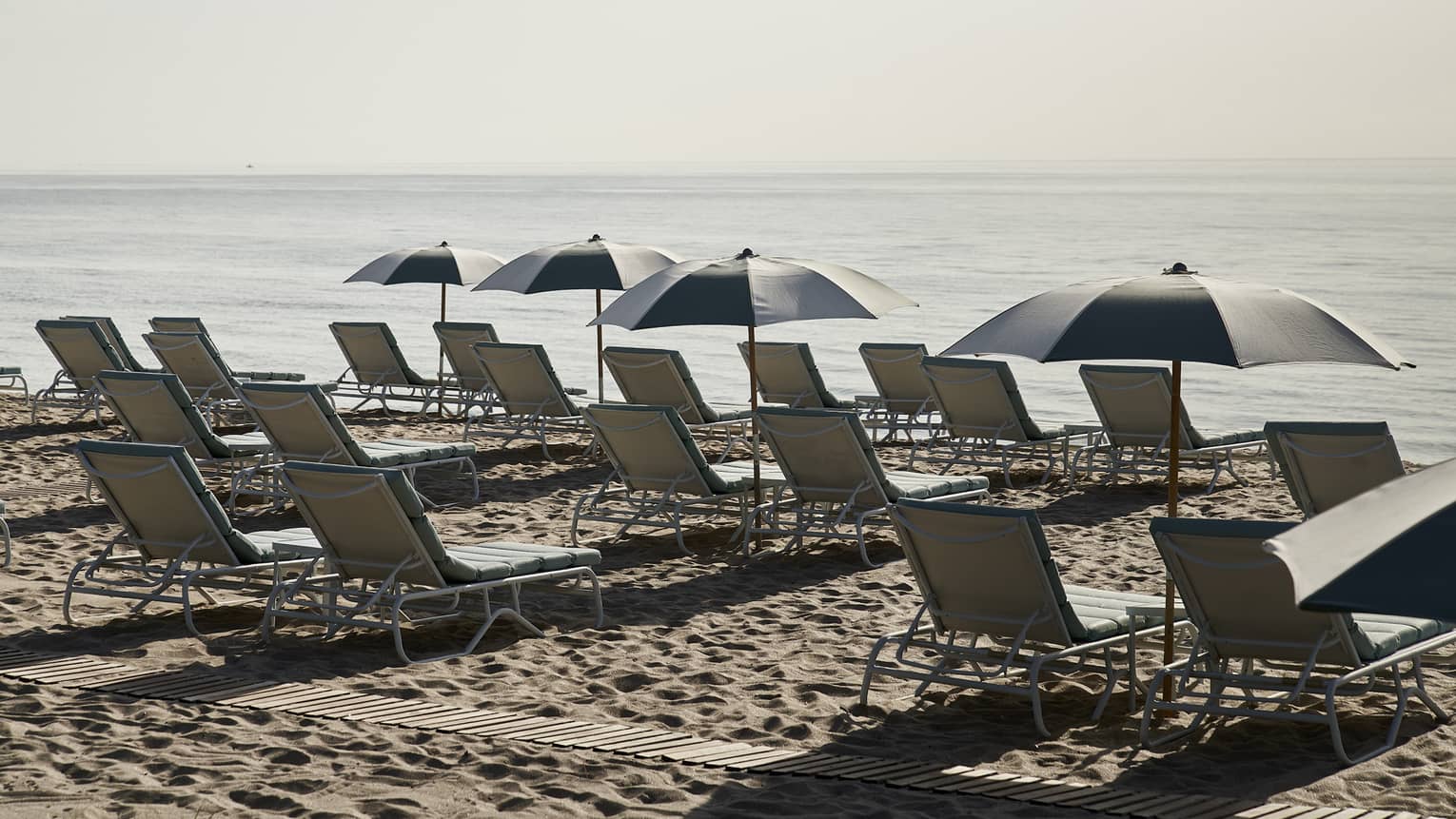 Lounge chairs and umbrellas line the sandy shore near the ocean, offering a perfect spot to relax and enjoy the serene seaside view.