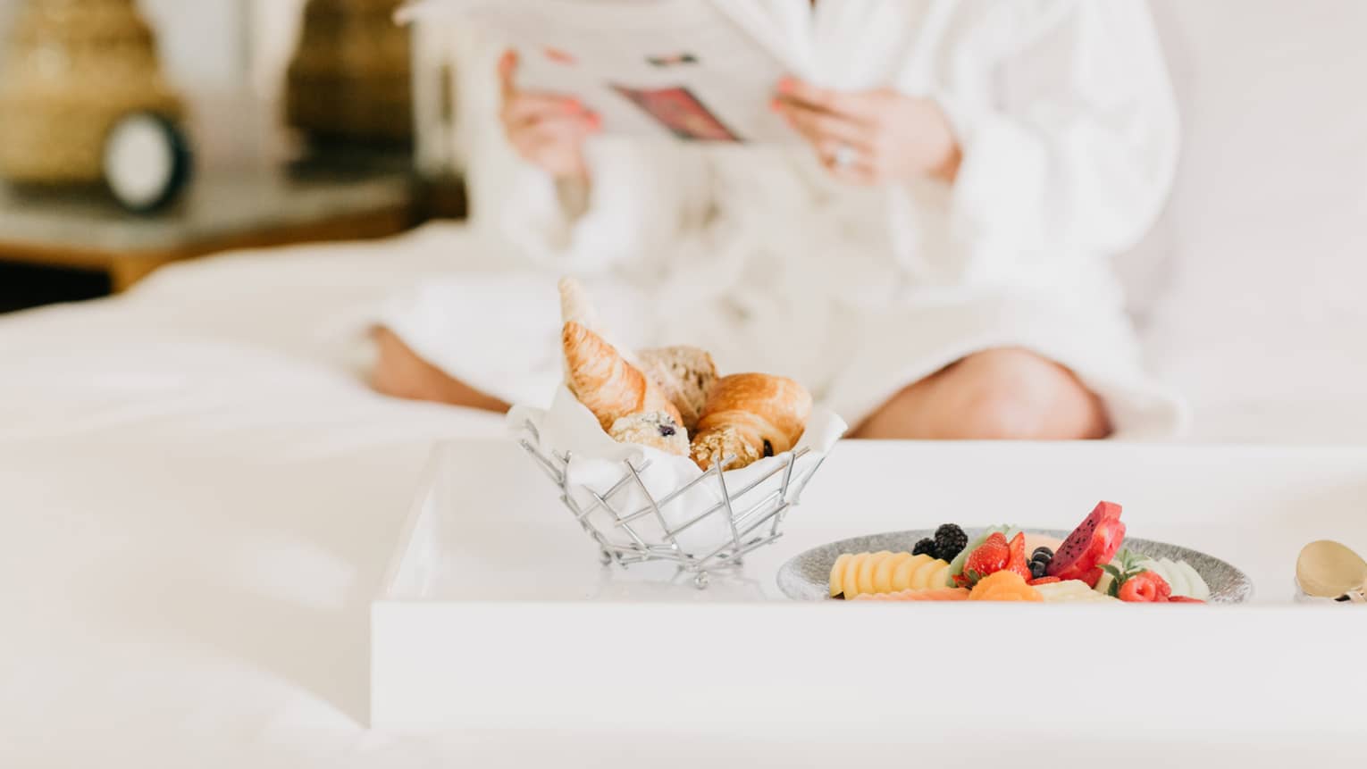 Pastries, fruit on in room dining tray, woman wearing white bath robe reads in background