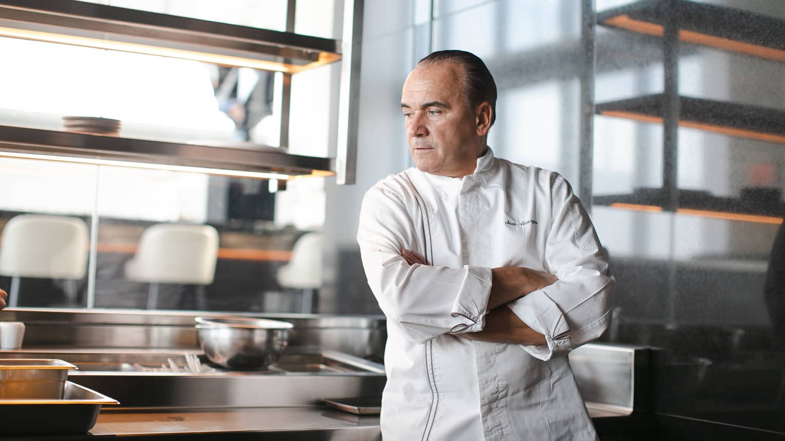 Chef Jean-Georges Vongerichten, wearing a white chef's jacket, leans against a kitchen workstation with his arms crossed