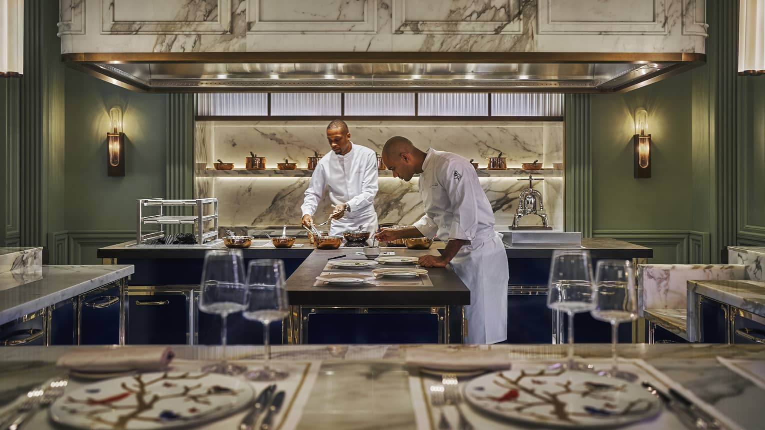 Two chefs work around a centre island in a green-walled open kitchen