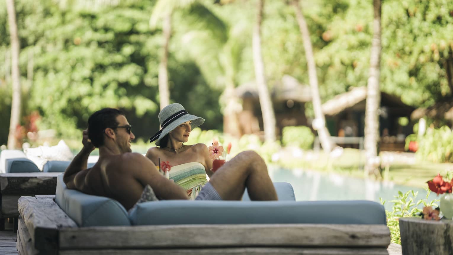 Couple lounging in swimwear on light-blue lounger poolside with palm trees in backdrop