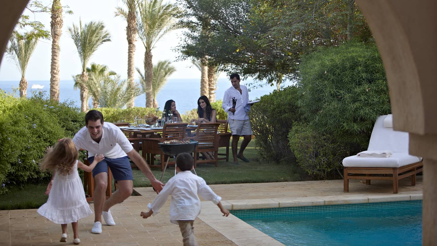 Man with open arms greets two children as they run toward him on patio deck near swimming pool